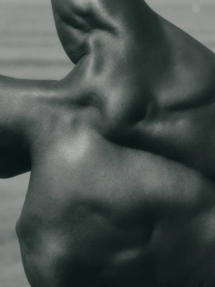 Close-up of a person's upper body and arm with smooth, dark skin, highlighting the shoulder, collarbone, and neck area.