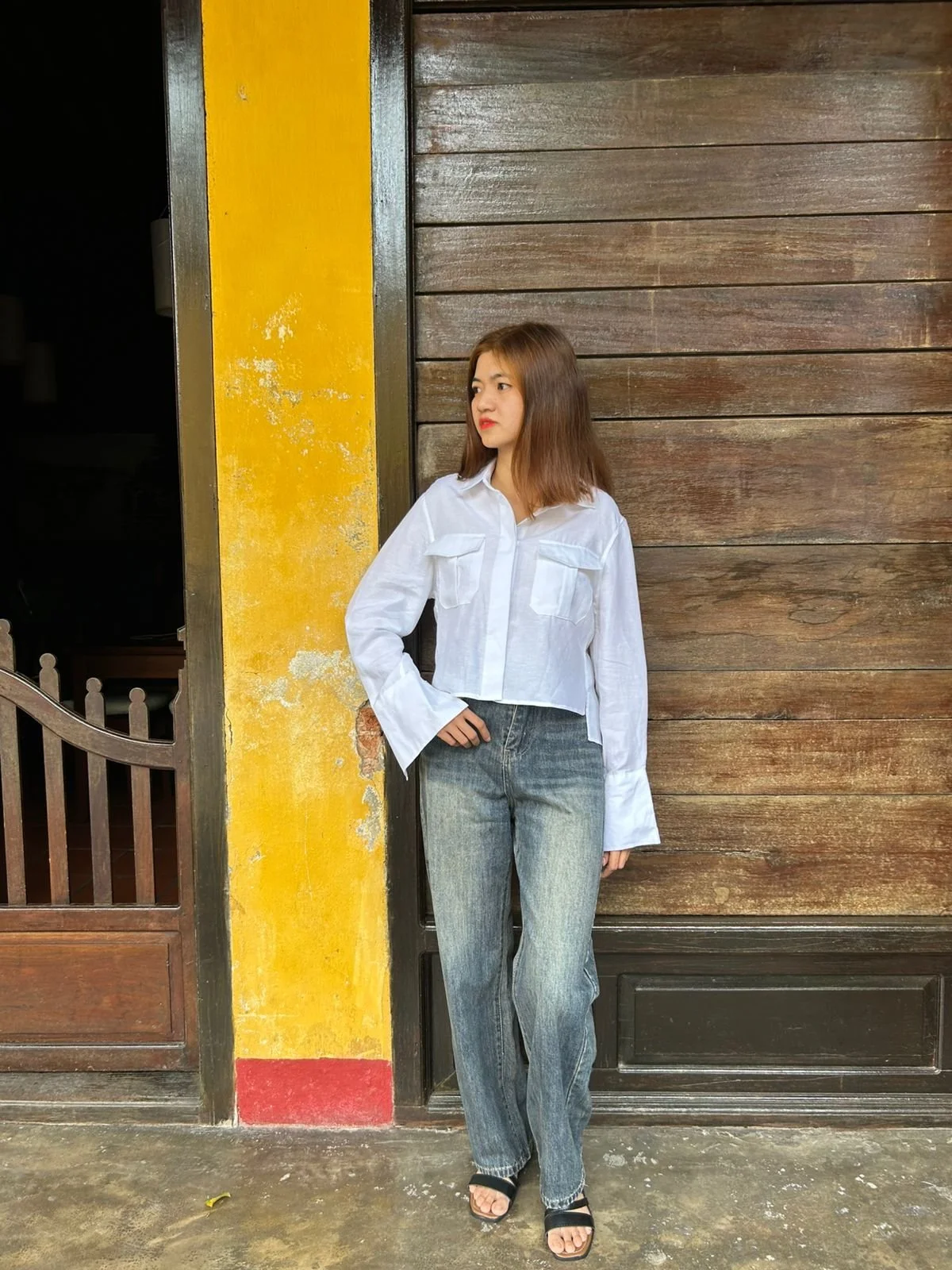 A woman in a white shirt and jeans standing against a yellow and wooden wall background.