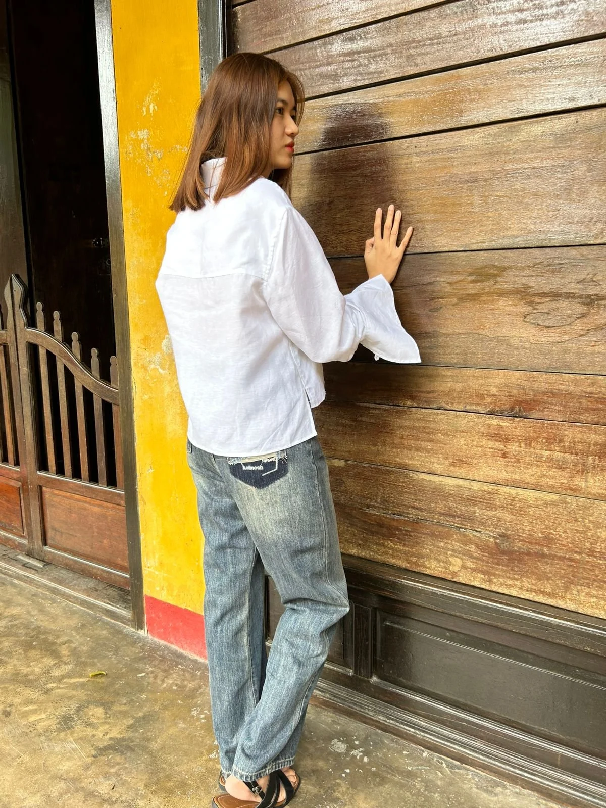 Person in a white shirt and jeans standing against a wooden wall, wearing sandals.