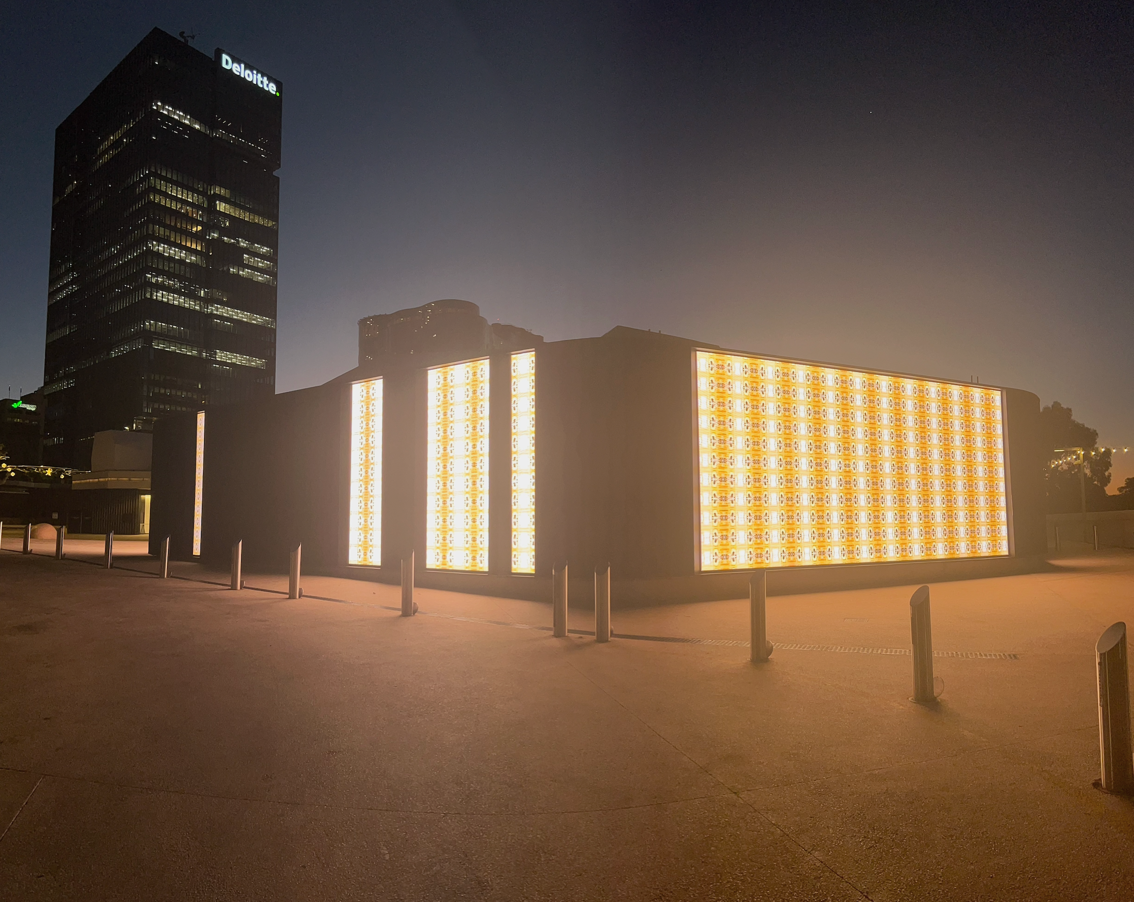 Nighttime cityscape with a tall Deloitte building and a lit-up structure in front, illuminated with orange and yellow patterned lights, surrounded by empty pavement and barriers.