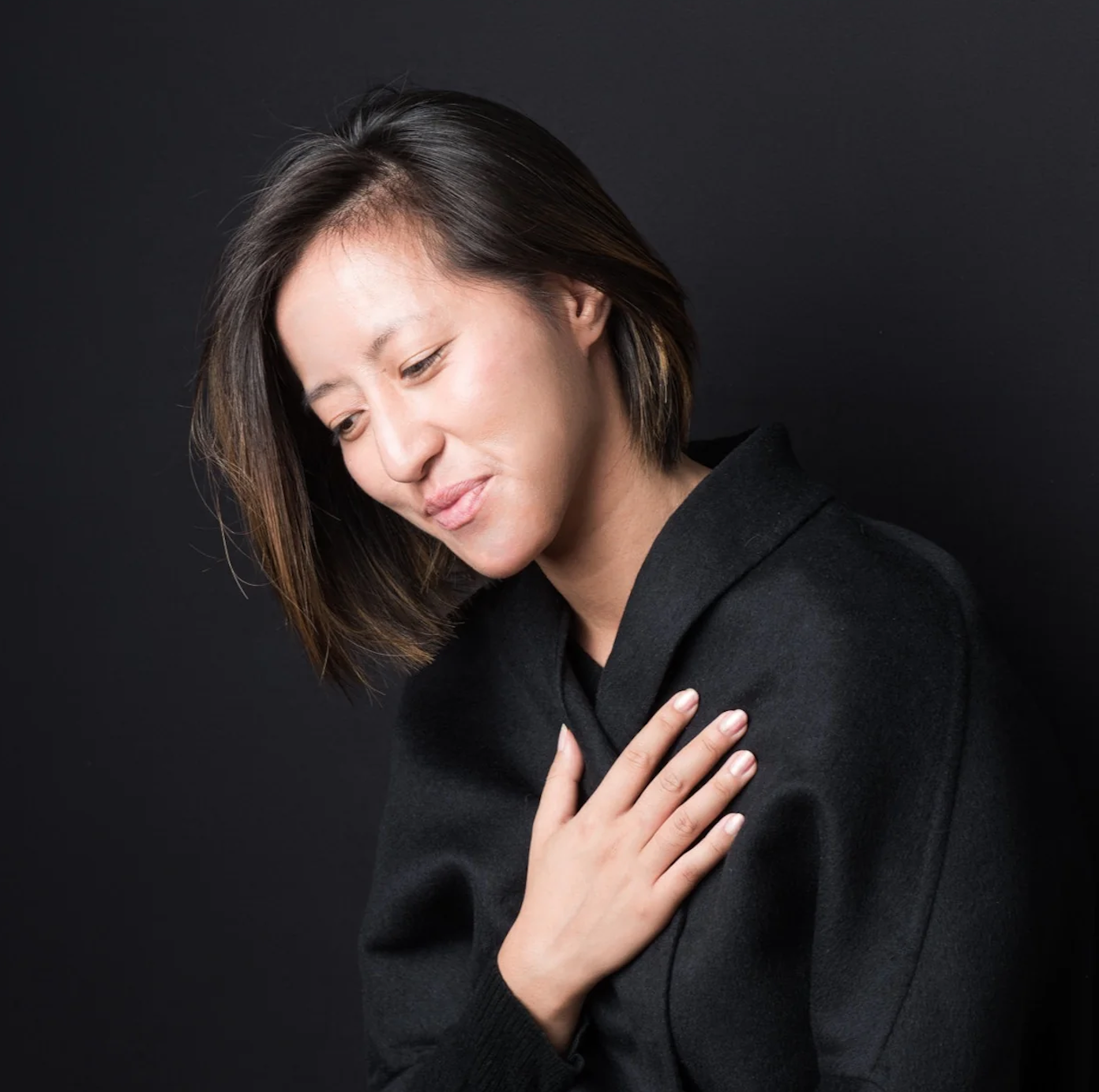 A woman with short dark hair and light skin, wearing a black sweater, is looking down with a gentle smile, her eyes closed, and her left hand placed on her chest against a black background.