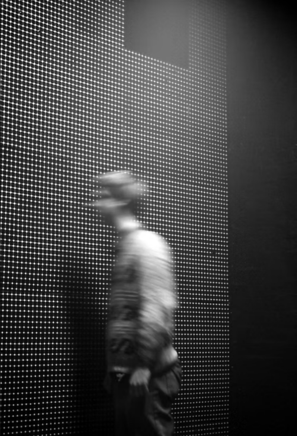Black and white photo of a person walking past a wall with illuminated dots creating a pixelated pattern.
