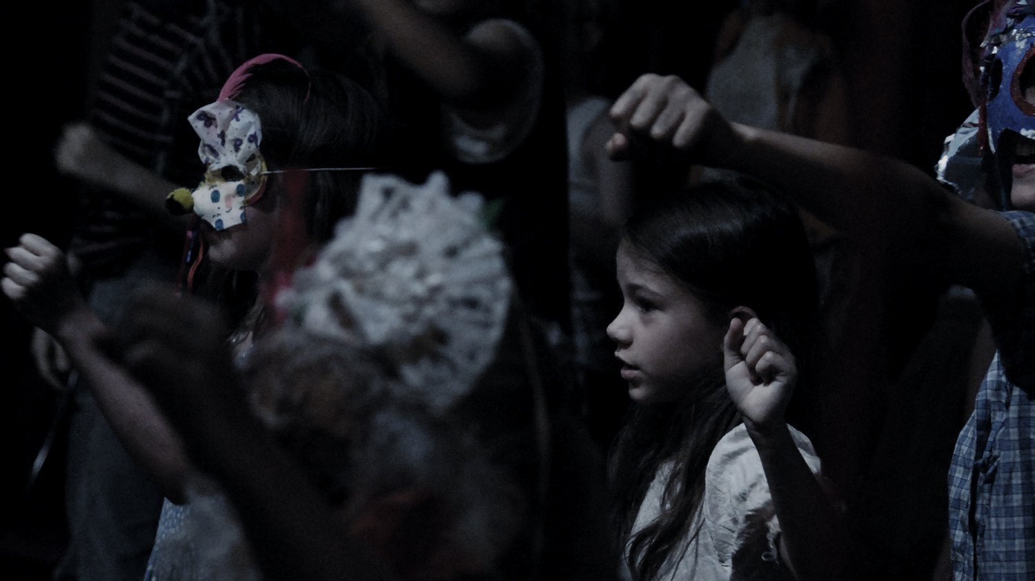 Children in a dark room, some wearing masks or face shields, appear to be praying or engaged in a religious activity.