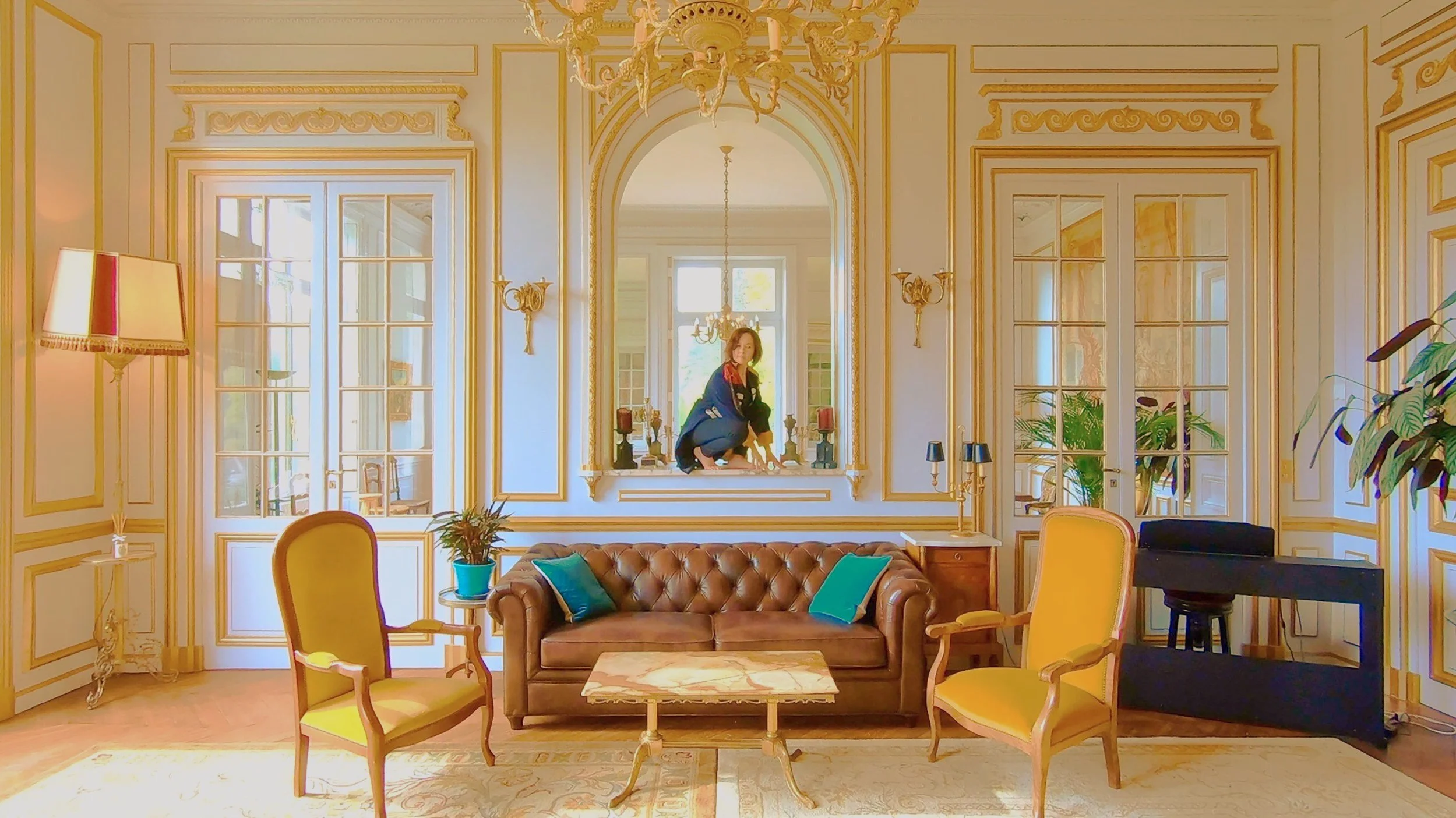 A woman crouching on a windowsill in a luxury room with ornate cream-colored walls and gold trim, surrounded by elegant furniture and decor, with sunlight streaming through large windows.