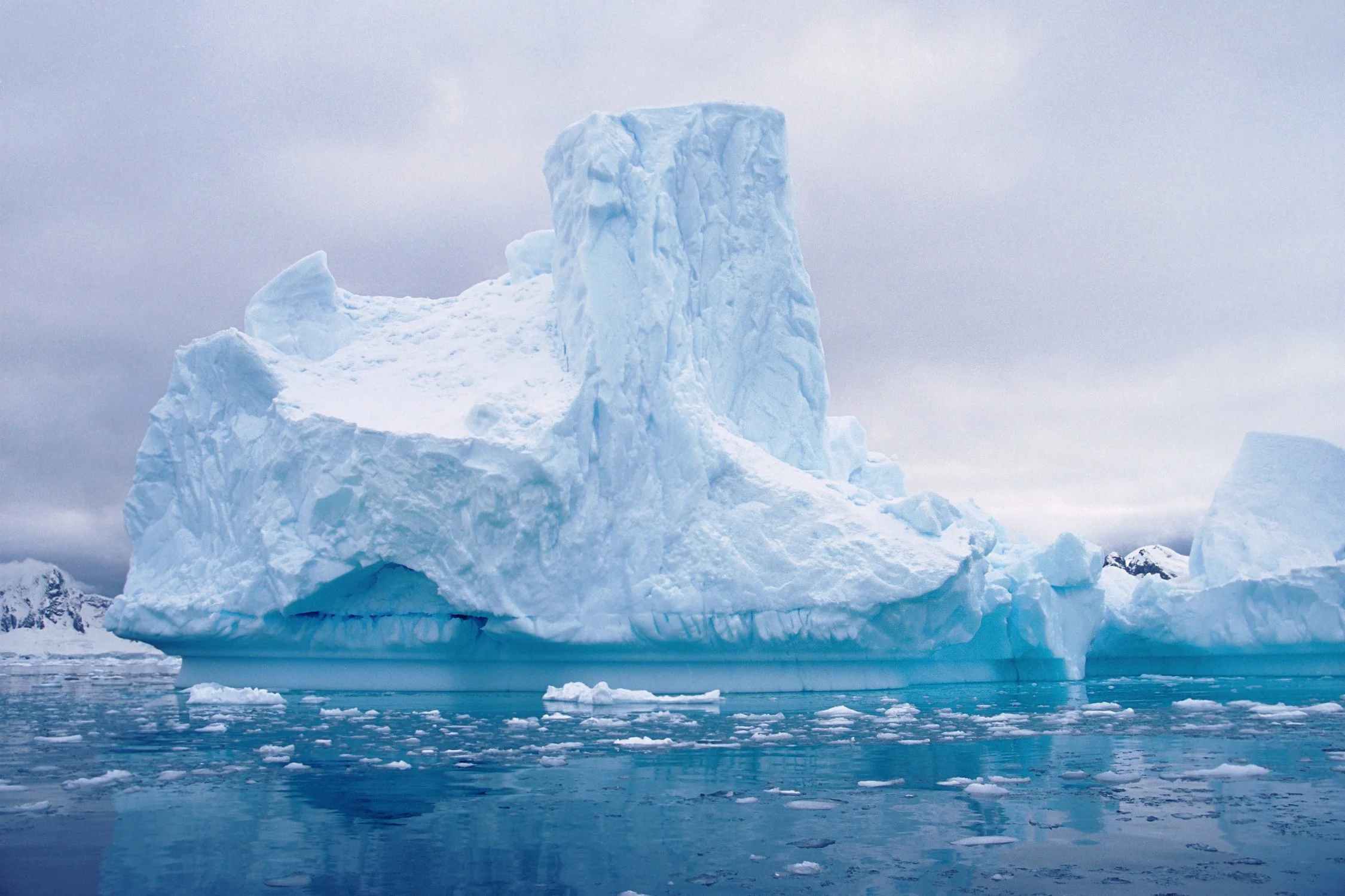 A large iceberg floating in icy blue water, with a smaller chunk nearby, under a cloudy sky.