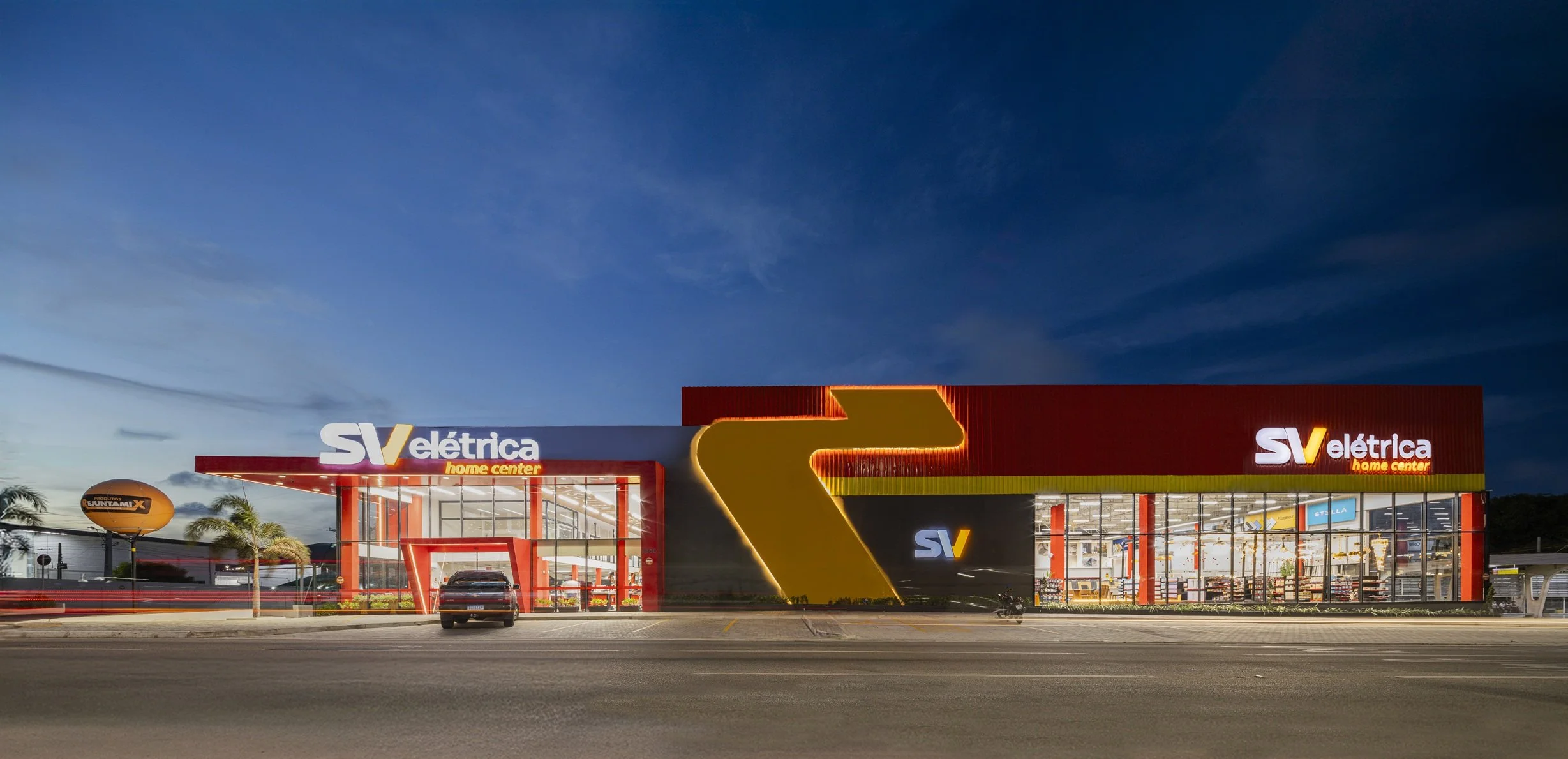 Fachada de uma loja de eletrônicos com iluminação noturna, sinalização luminosa de 'SV elétrica home center' e ambiente interno visível através de grandes janelas.