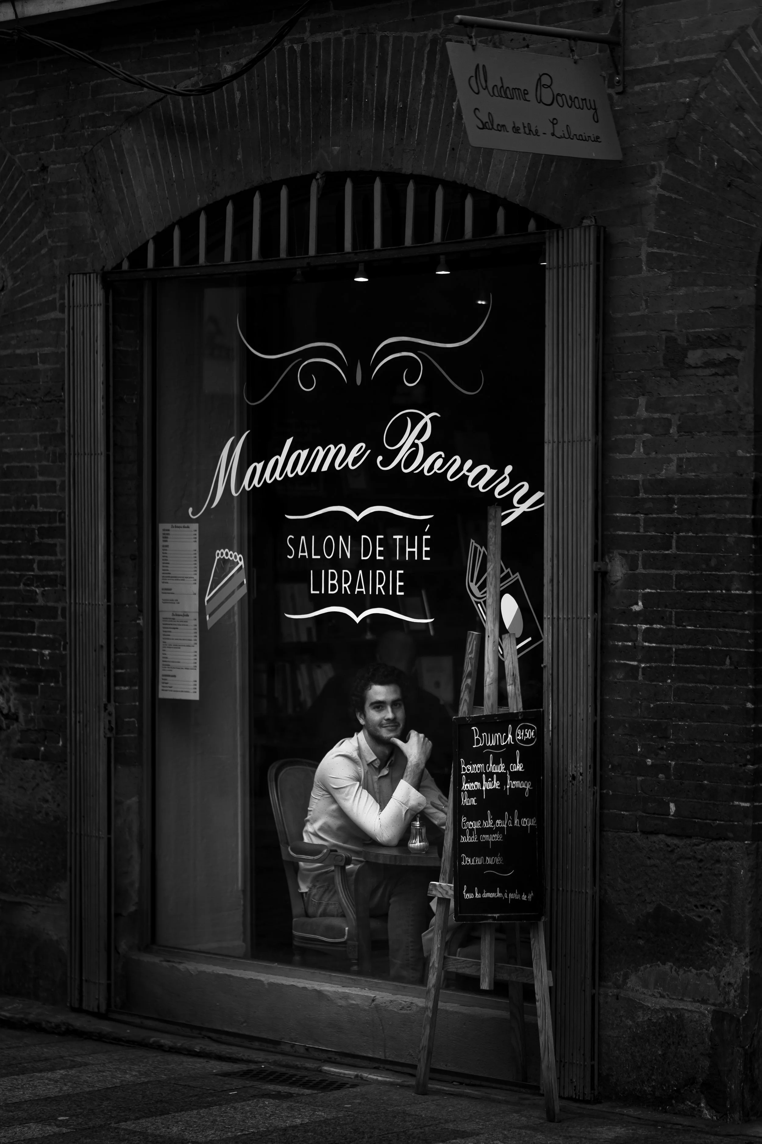 Une rue avec une vitrine de salon de thé nommé Madame Bovary, avec un homme assis à une table à l'intérieur, visible à travers la vitre. Le panneau indique un menu ou une offre spéciale.
