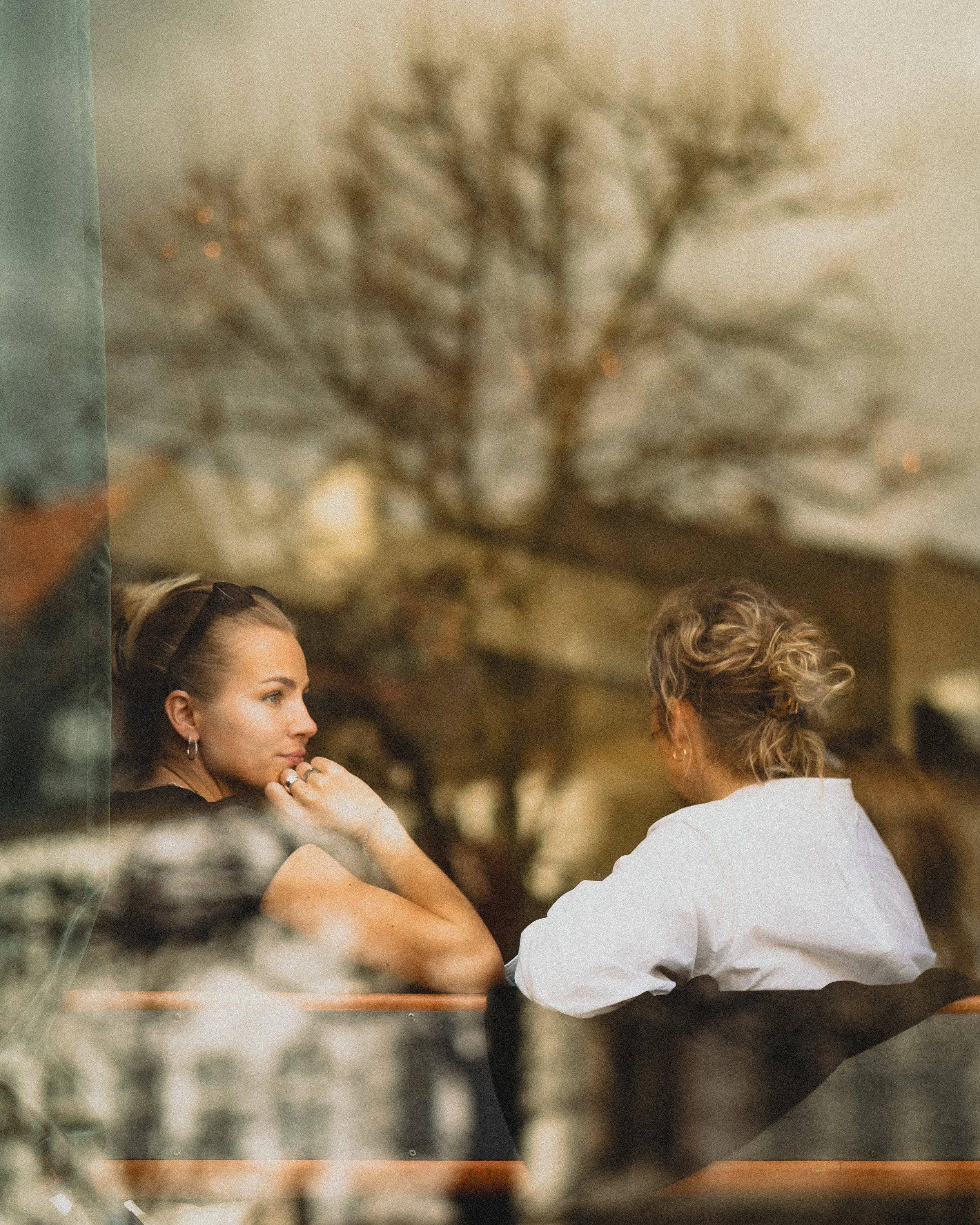 Deux femmes assises à une table dans un café, vues à travers une vitre, conversation intime, arrière-plan flou de branches d'arbres.
