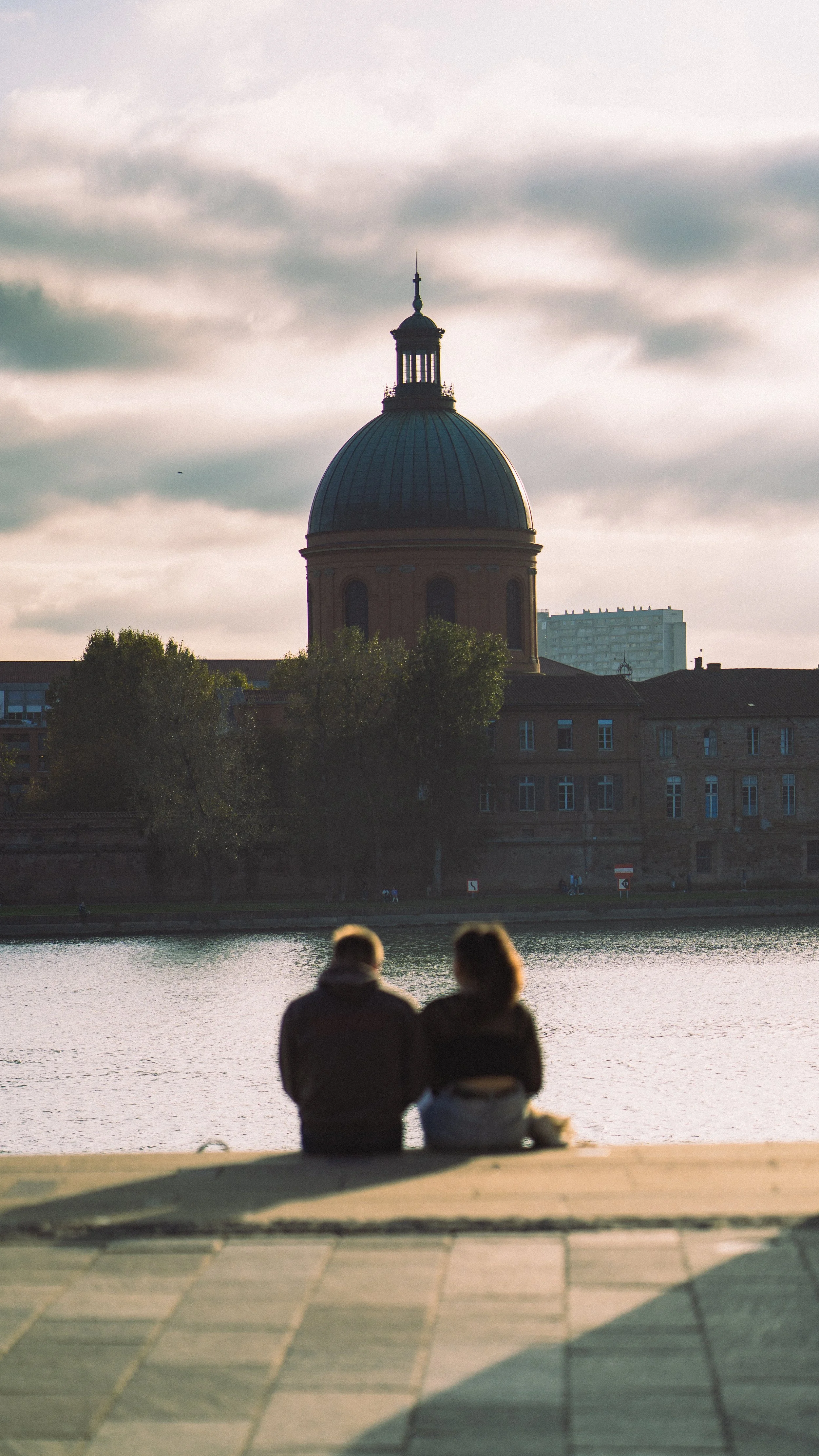 Deux personnes assises près de l'eau, face à un bâtiment avec un grand dôme, au coucher du soleil.