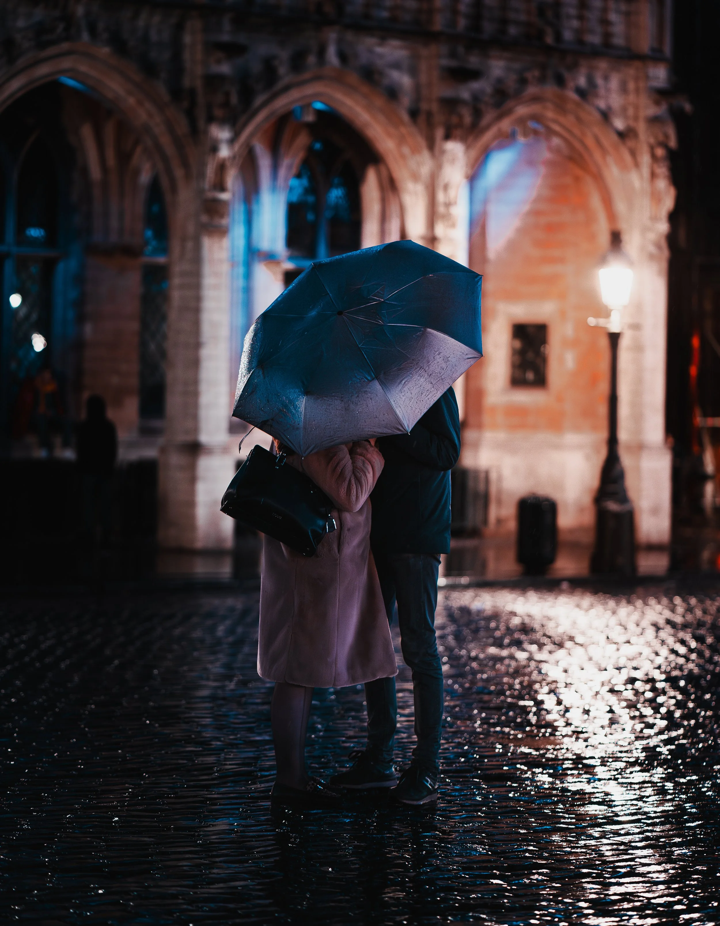 Deux personnes sous un parapluie dans une rue pavée mouillée, avec un ancien bâtiment illuminé en fond, lors d'une nuit pluvieuse.