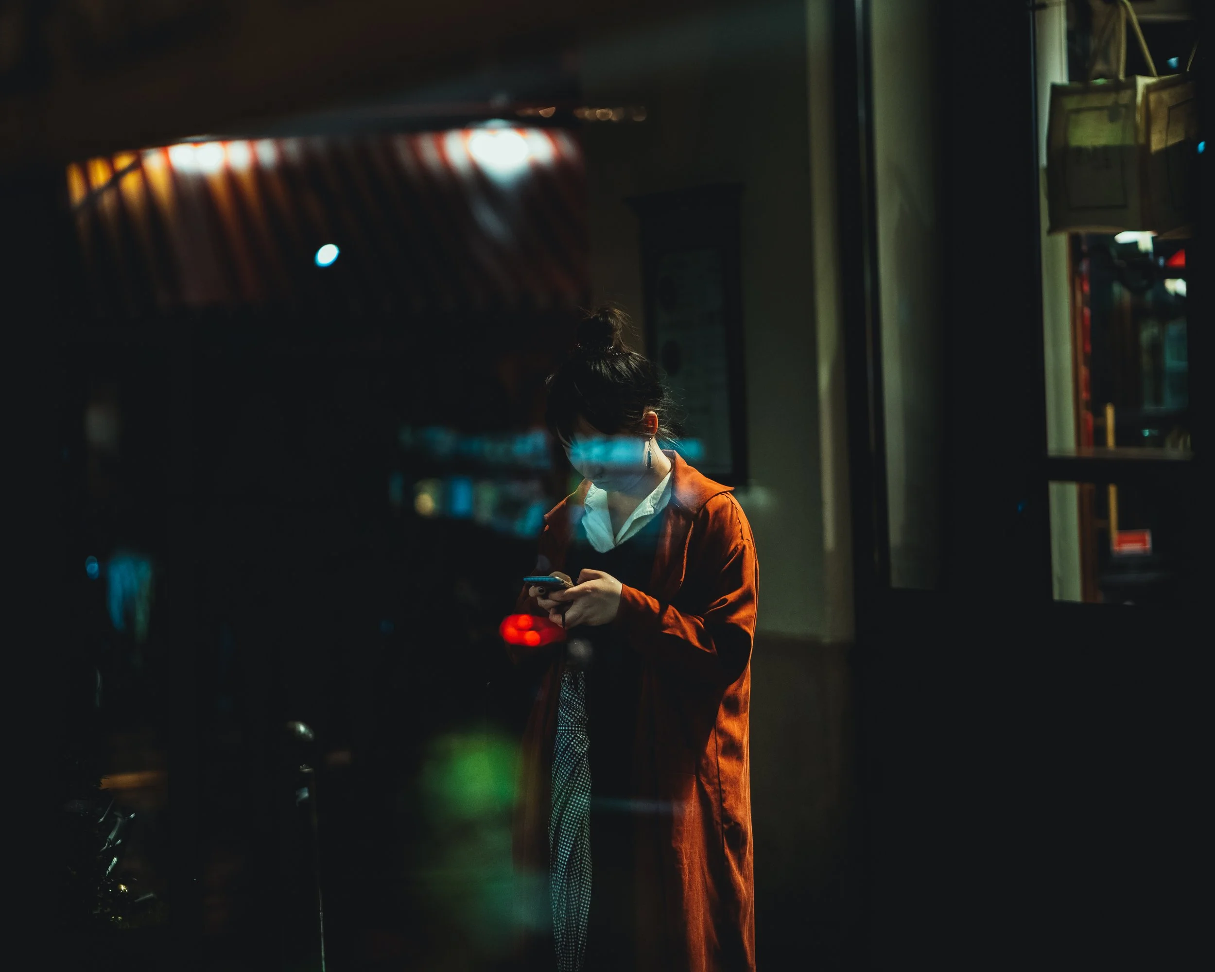 Femme portant un manteau orange foncé et un masque de protection, regardant son téléphone dans un environnement urbain la nuit.
