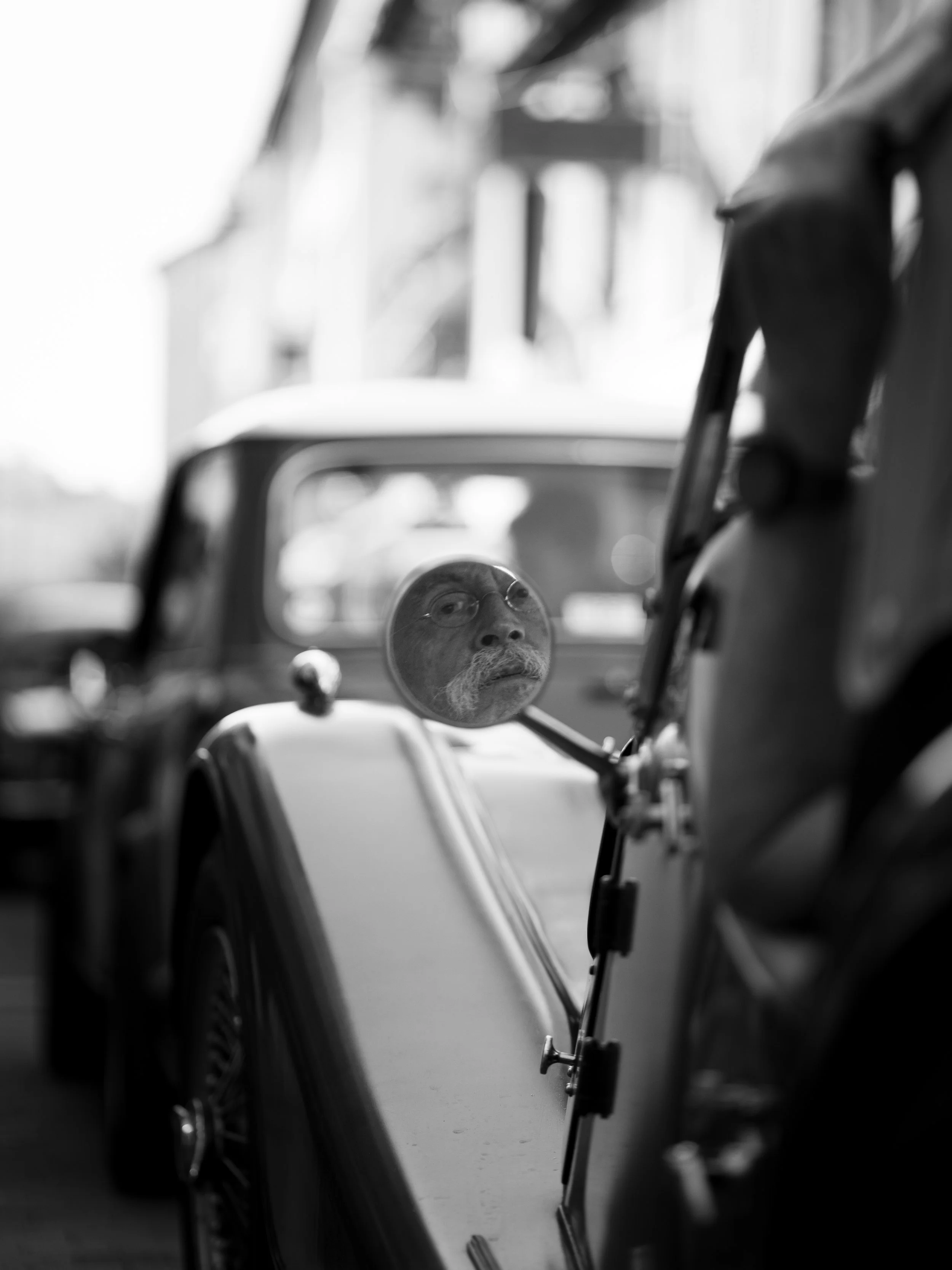Un miroir rond fixé à une vieille voiture montre le visage d'un homme avec des lunettes et une moustache blanche. La voiture est en noir et blanc, avec un environnement flou en arrière-plan.