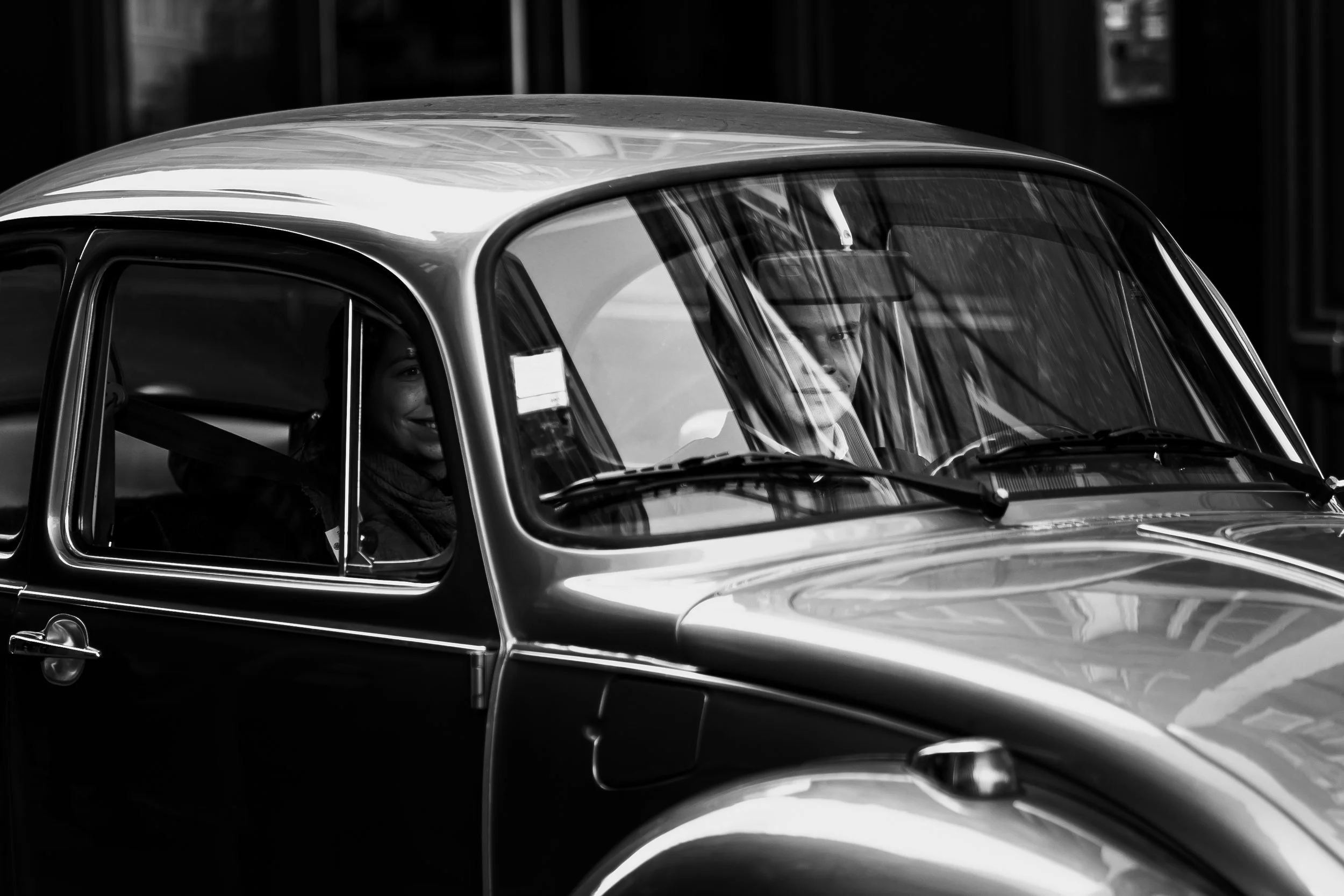 Deux personnes souriantes assises dans une voiture ancienne, vue de côté, en noir et blanc.