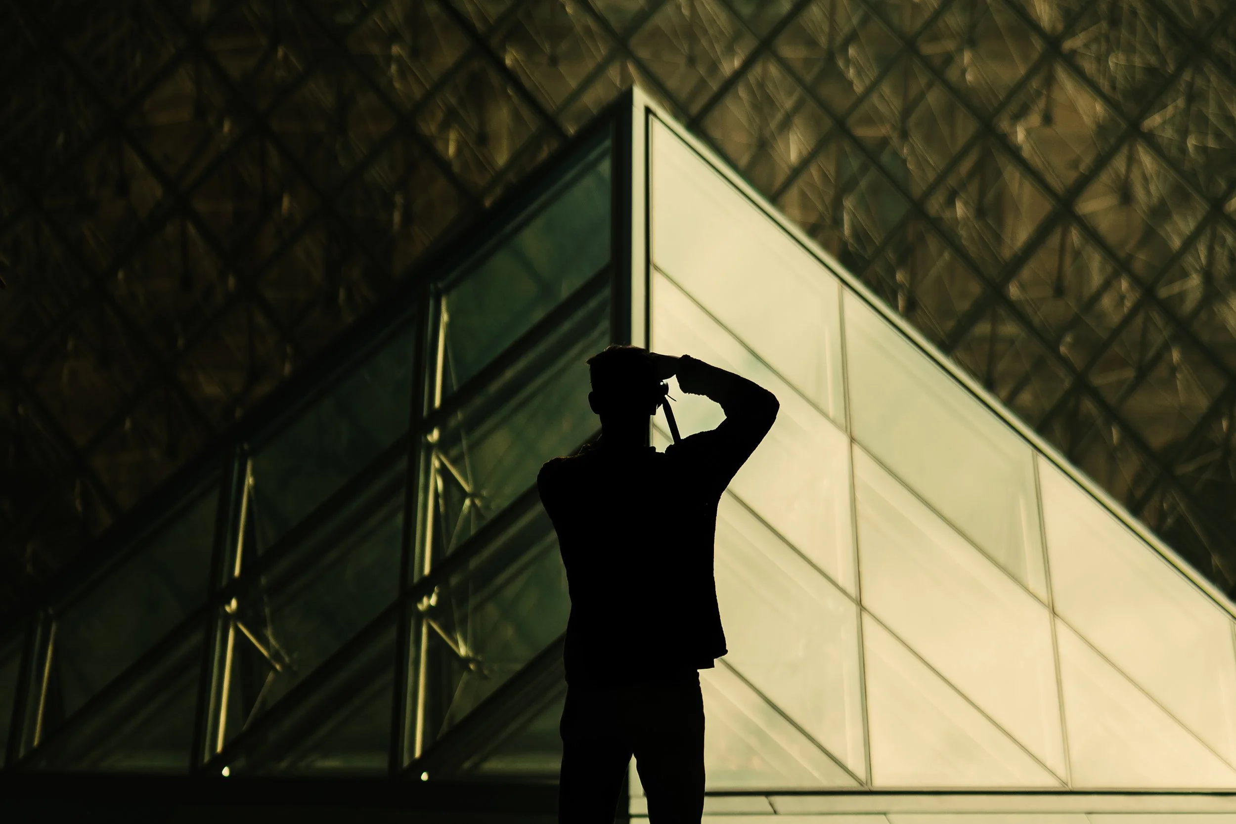 Silhouette d'une personne prenant une photo devant un bâtiment moderne en verre, la scène est sombre, probablement prise à l'extérieur la nuit.
