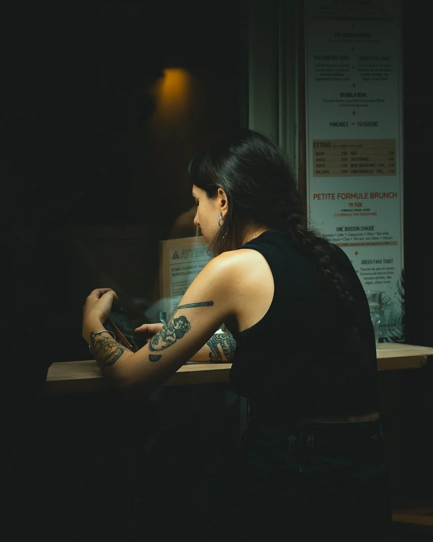 Jeune femme avec des tatouages assise à un comptoir dans un restaurant ou un café, regardant son téléphone, dans un environnement sombre.