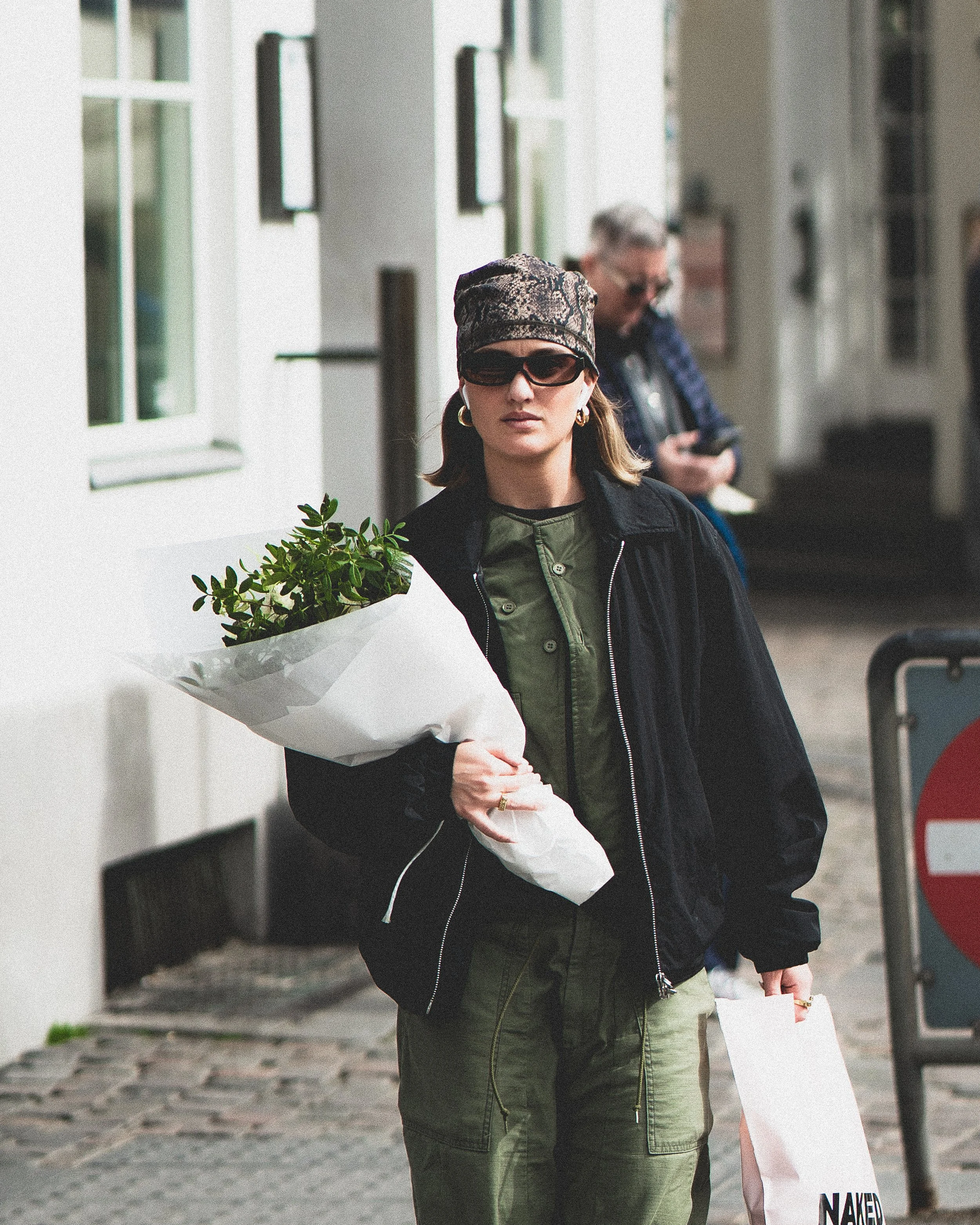 Une femme portant un chapeau, des lunettes de soleil, une veste noire, une chemise verte, et un pantalon vert, tenant un bouquet de fleurs blanches, en marchant dans une ville. En arrière-plan, un homme utilisant son téléphone.