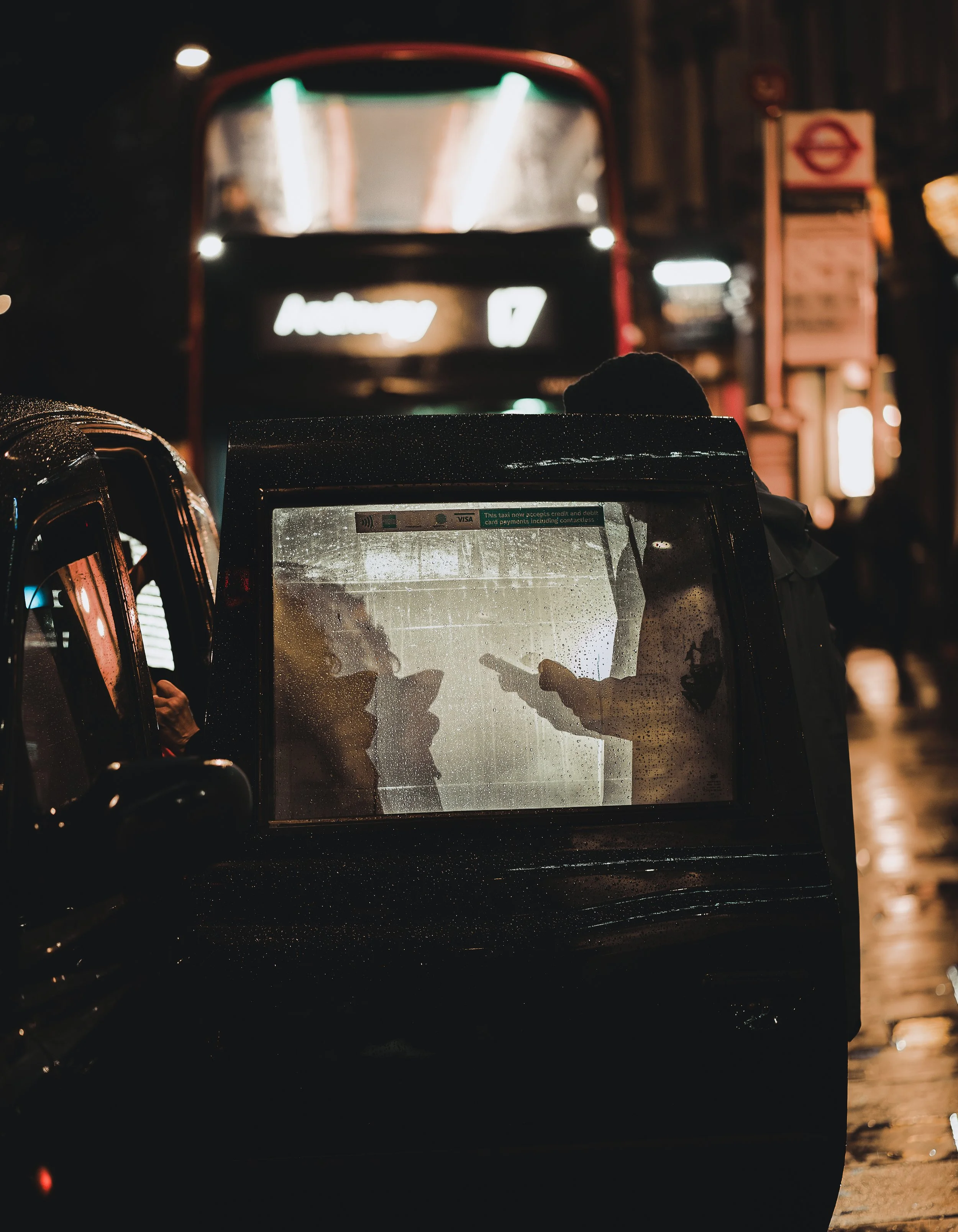 Deux personnes utilisant des téléphones portables dans une voiture noire sous la pluie, avec un autobus en arrière-plan dans une scène urbaine nocturne