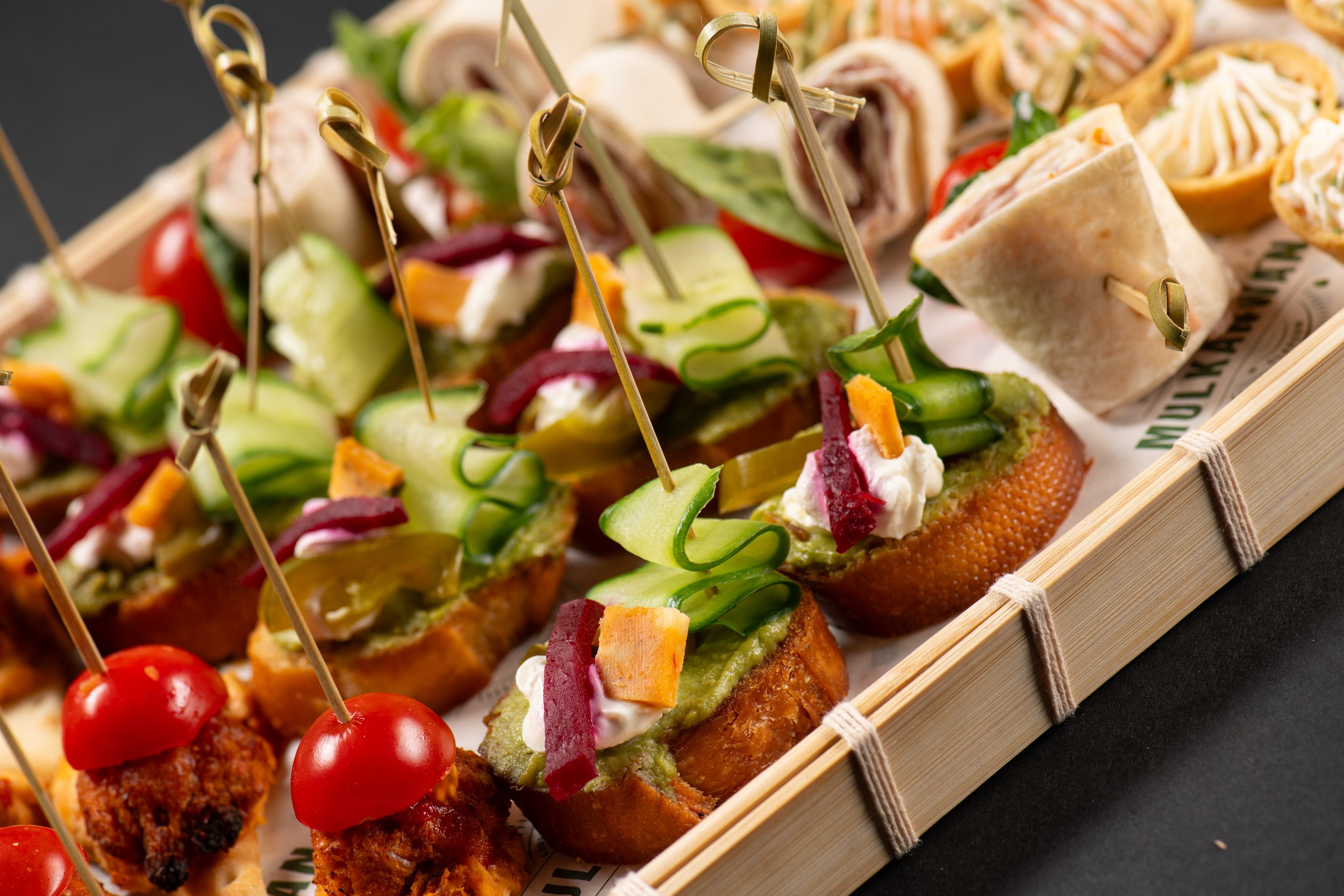 A tray of assorted appetizers featuring bruschetta with toppings like cucumber and cream, cherry tomatoes on skewers, and sandwich pinwheels.