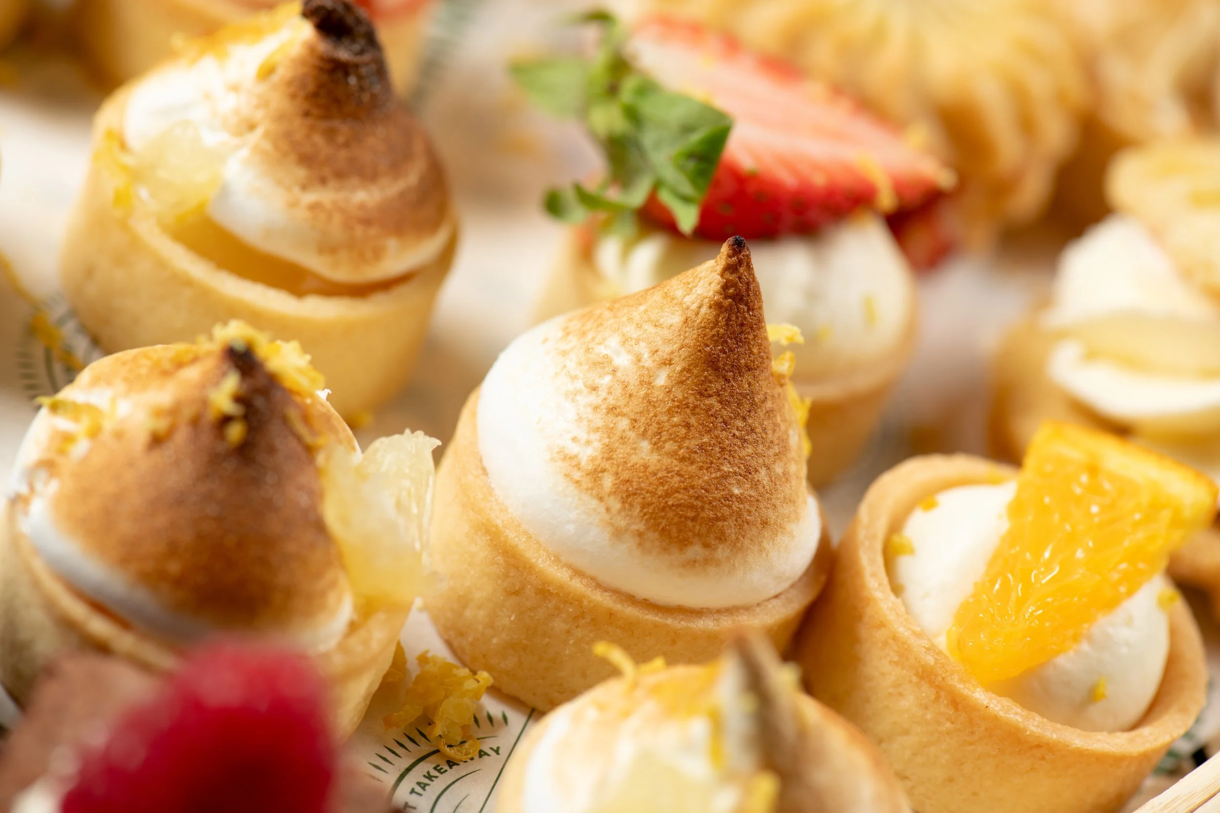 Close-up of assorted mini desserts with meringue topping, fresh strawberries, and orange slices, presented on a tray.