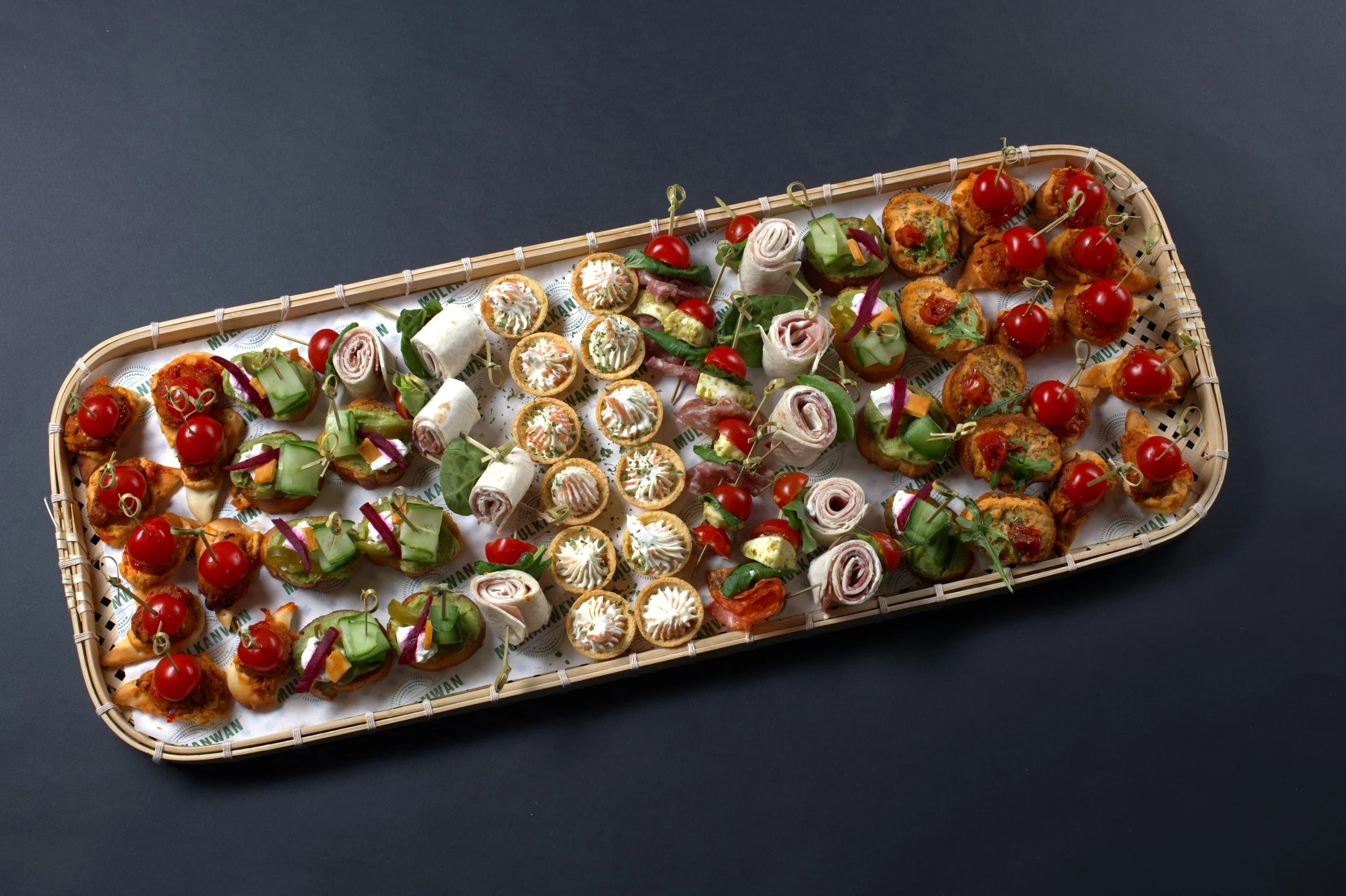 Assorted appetizer platter with canapés, wraps, tartlets, and skewers featuring cherry tomatoes, cucumbers, and deli meats on a bamboo tray.