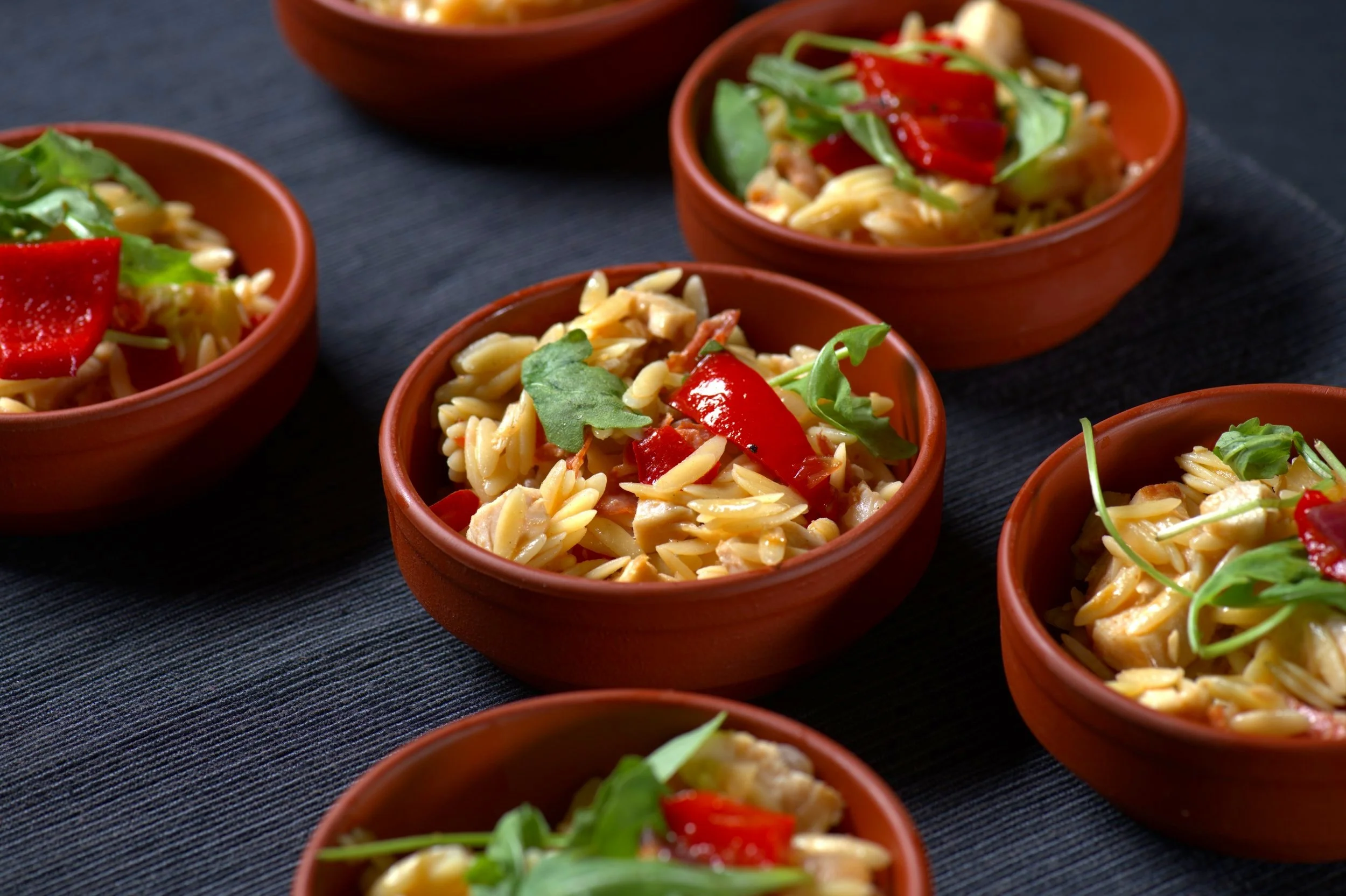 Small terracotta bowls with orzo pasta, red bell peppers, and arugula on a dark background.