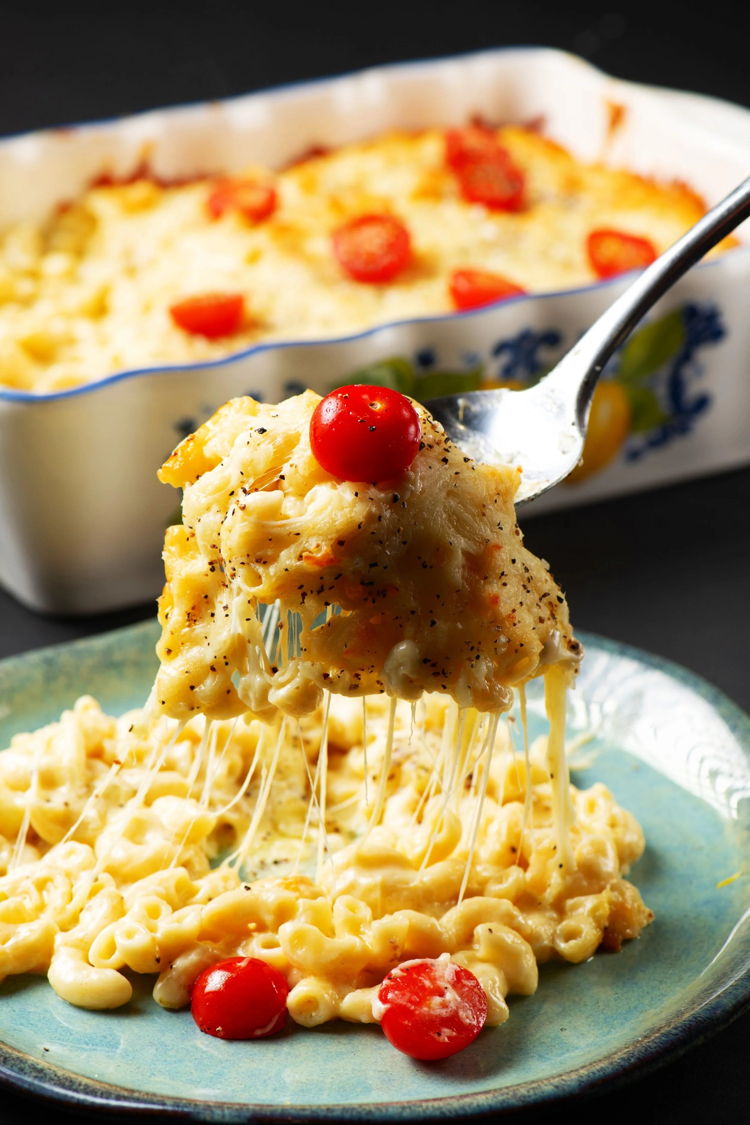 Cheesy baked macaroni with cherry tomatoes on a plate, with fork lifting a serving from a casserole dish in the background.