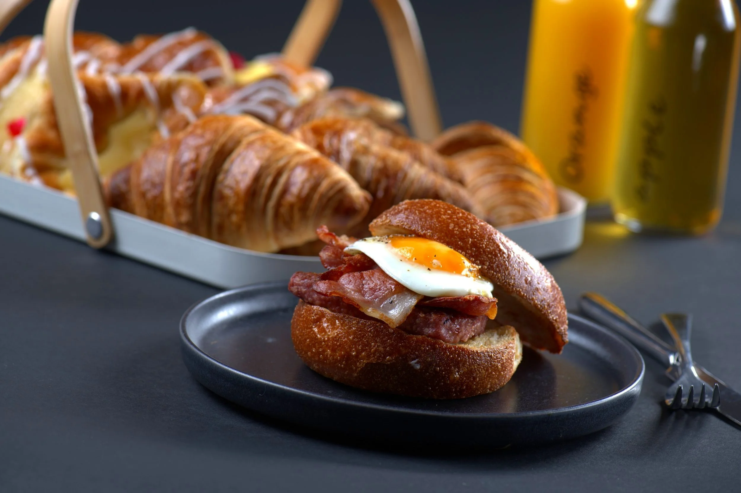 Breakfast sandwich with egg and bacon on a black plate, croissants in a tray, and bottles of orange and apple juice in the background.