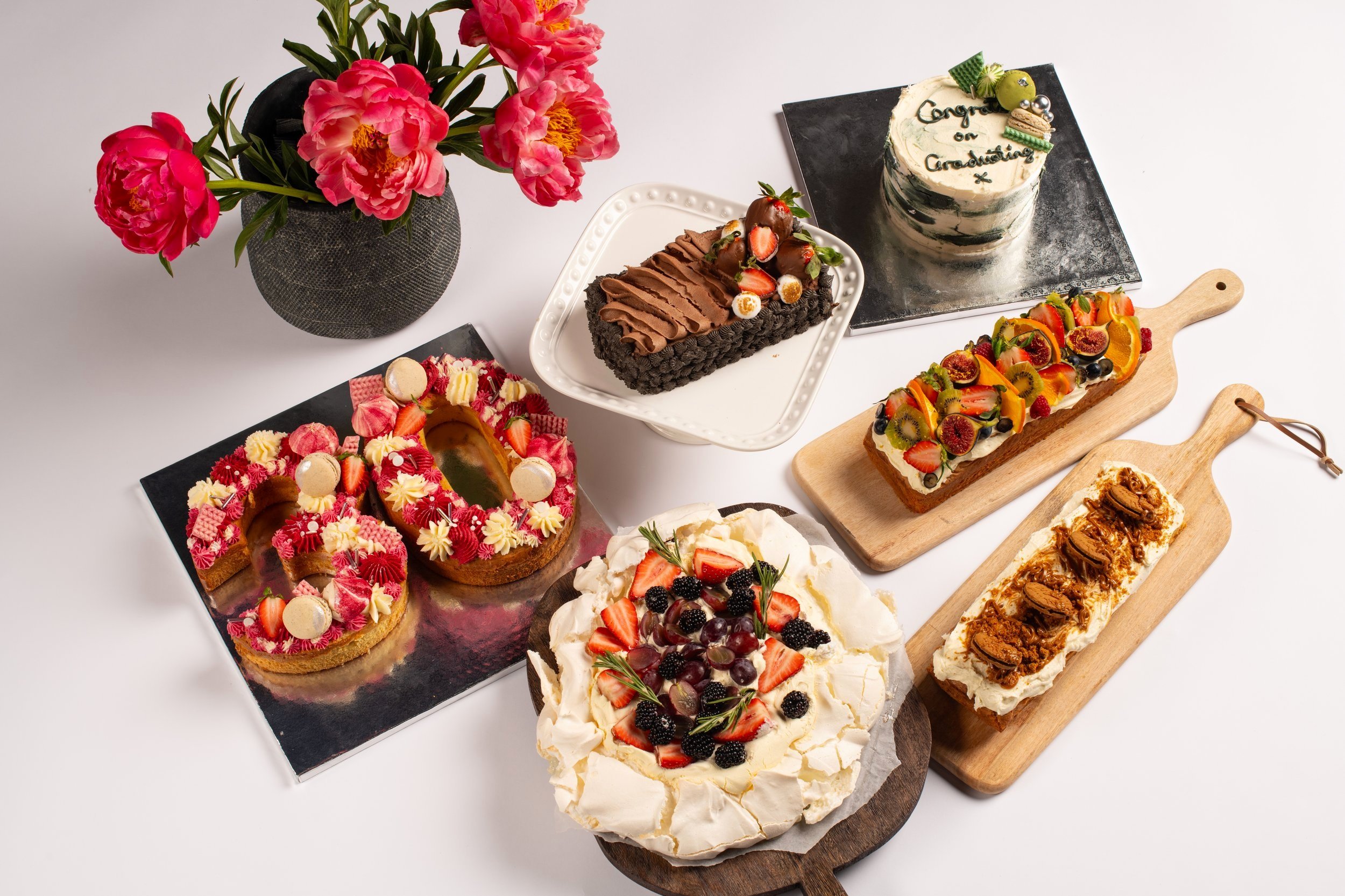 Variety of decorated cakes and desserts on a table with a vase of pink flowers.