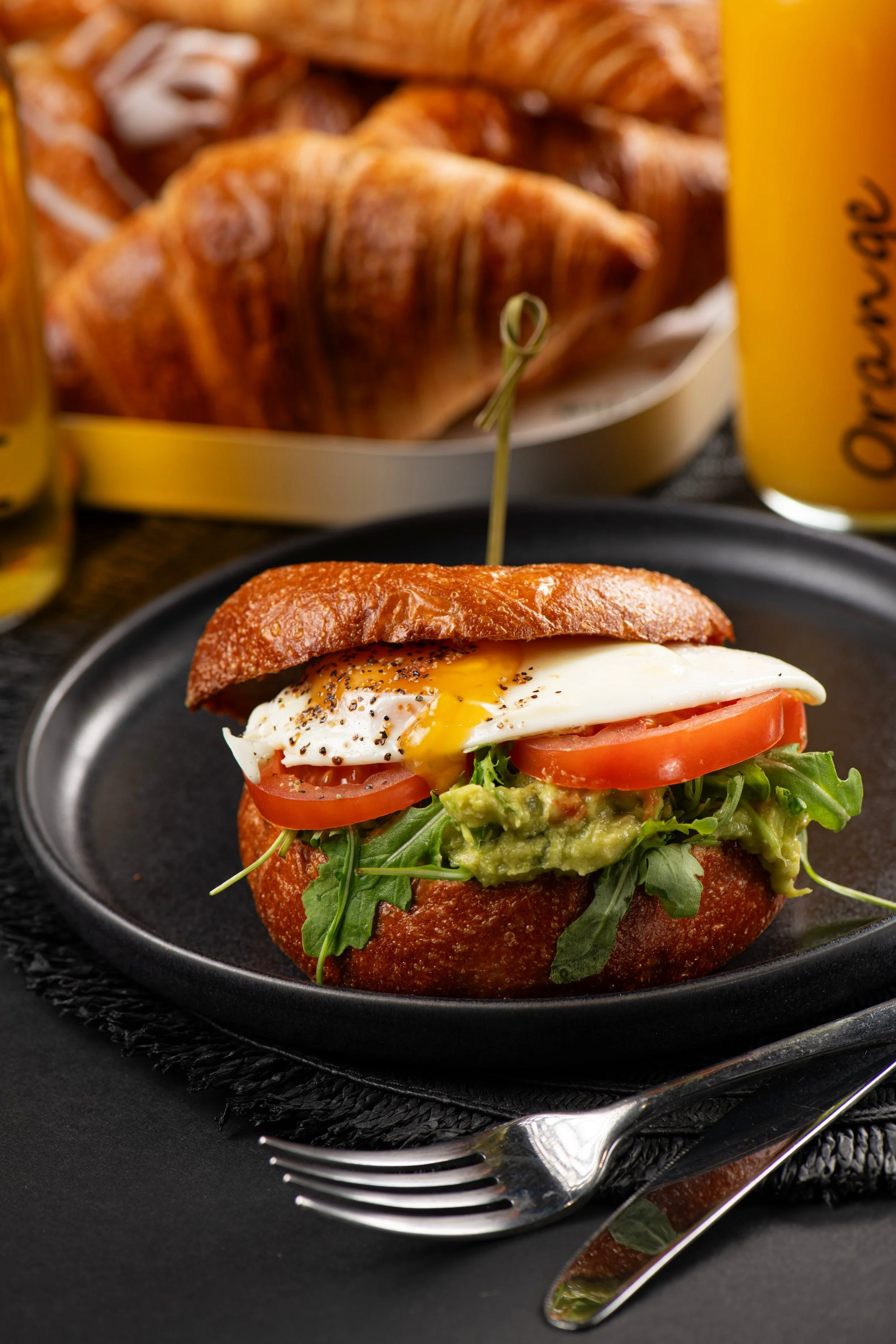Breakfast sandwich on a plate with a runny egg, tomato slices, arugula, and avocado on a brioche bun, surrounded by croissants and a glass of orange juice.