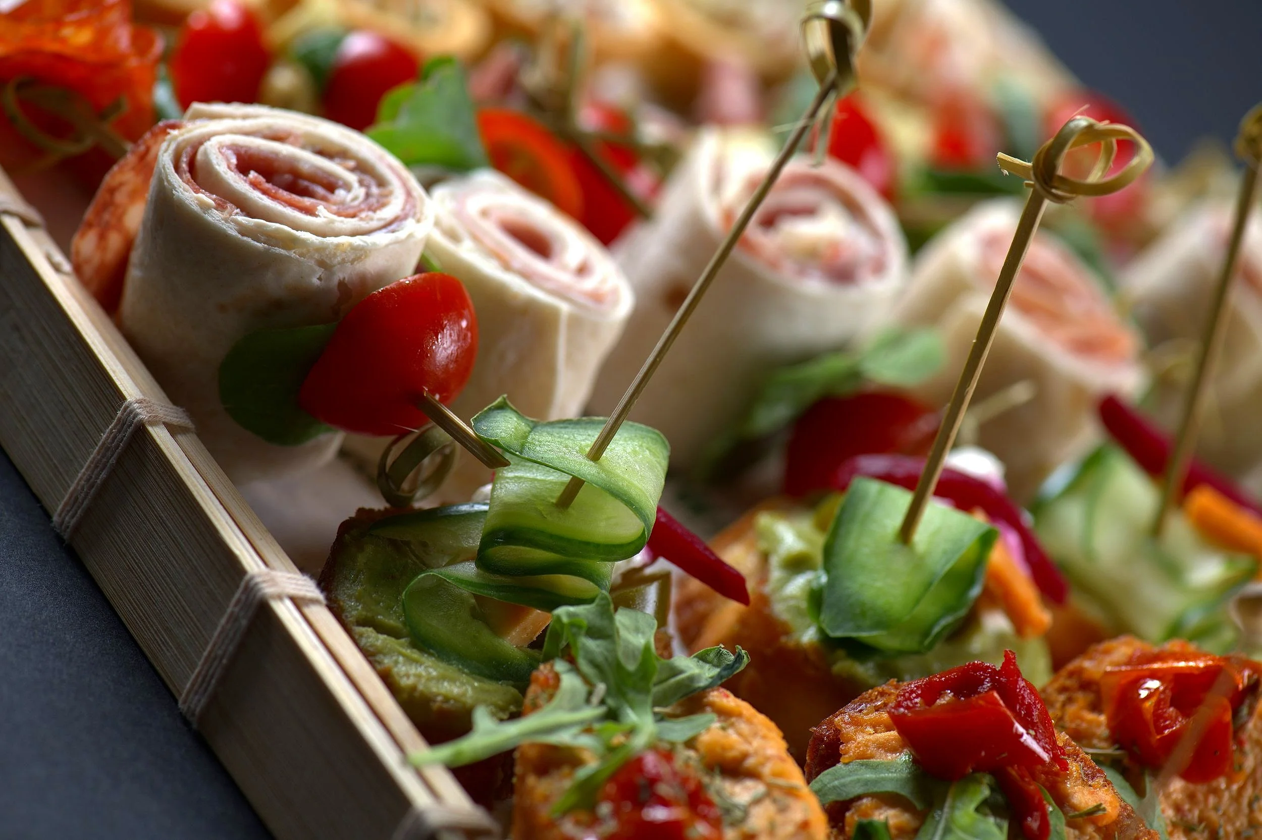 Assorted appetizers with wrapped tortilla rolls, cherry tomatoes, cucumber slices, and skewers.
