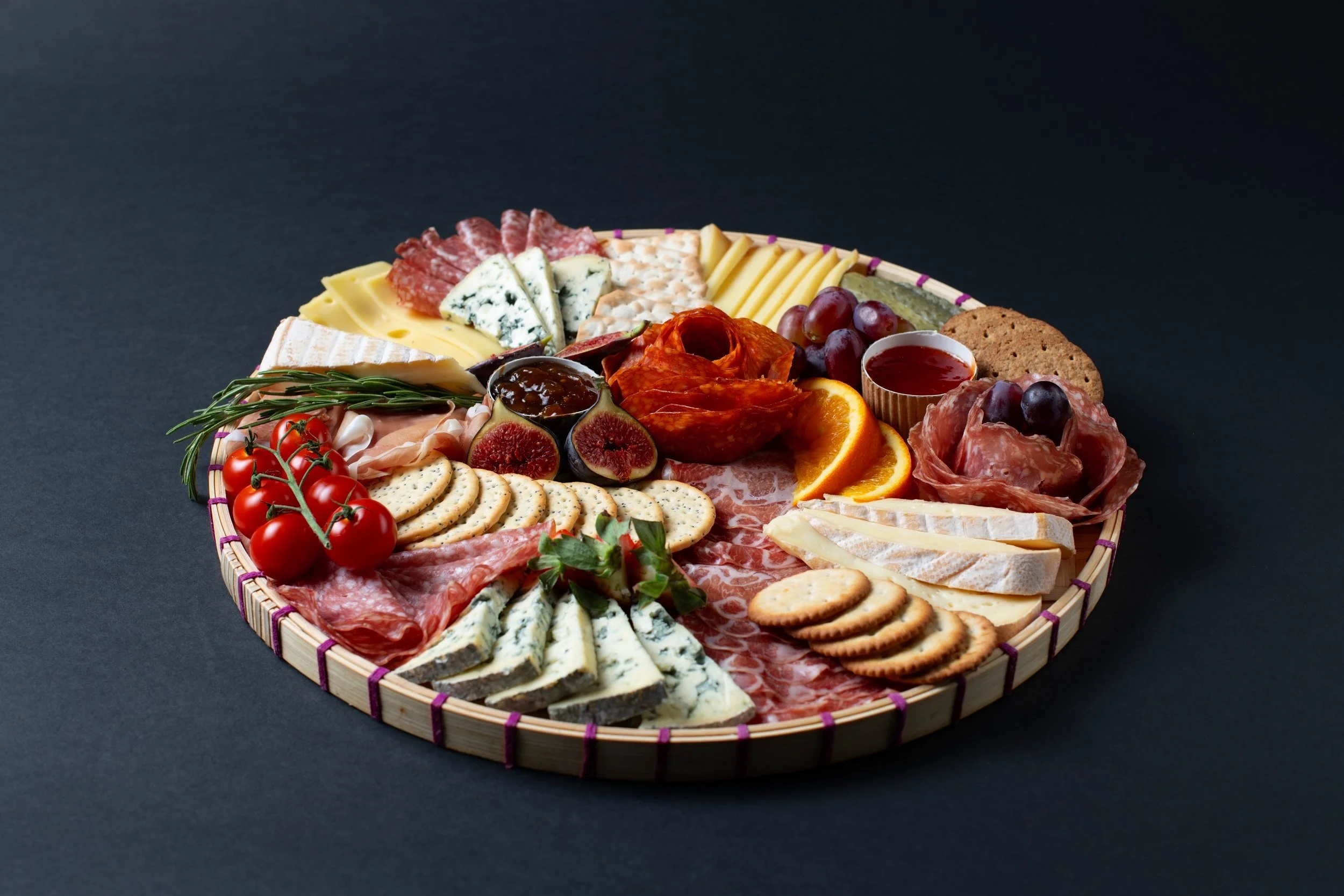 Charcuterie board with assorted cheeses, meats, crackers, grapes, figs, cherry tomatoes, orange slices, and jam on a round wooden platter.