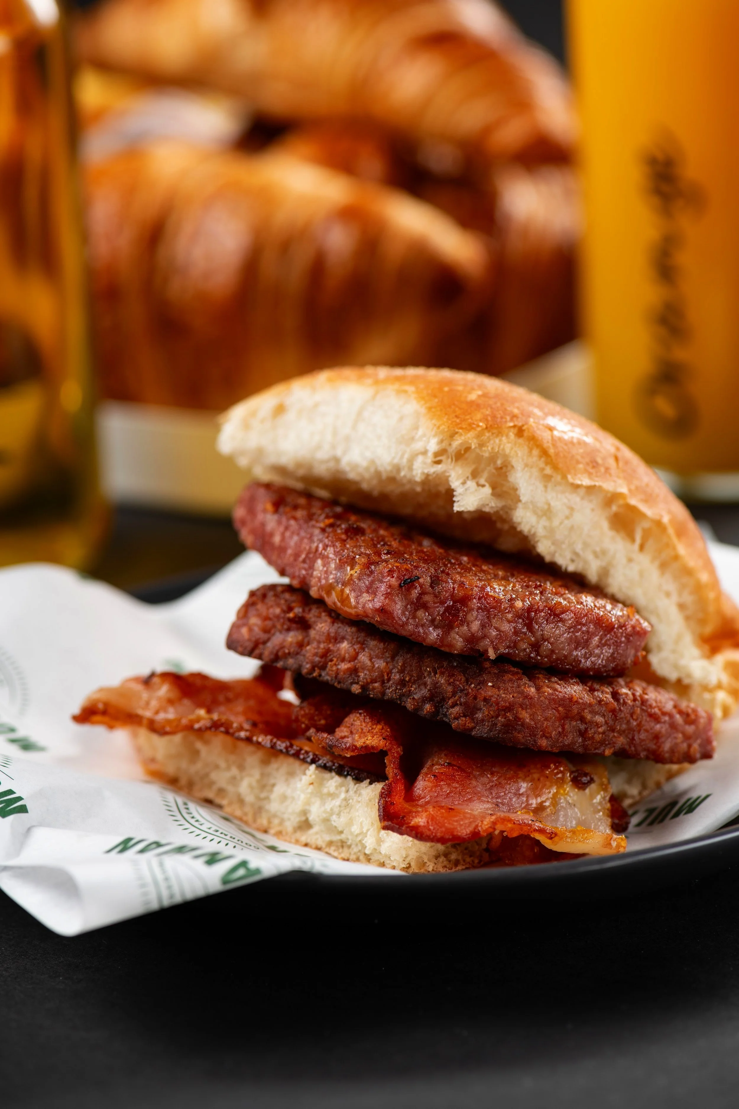 Close-up of a bacon and sausage breakfast sandwich on a bun, with croissants and orange juice in the background.