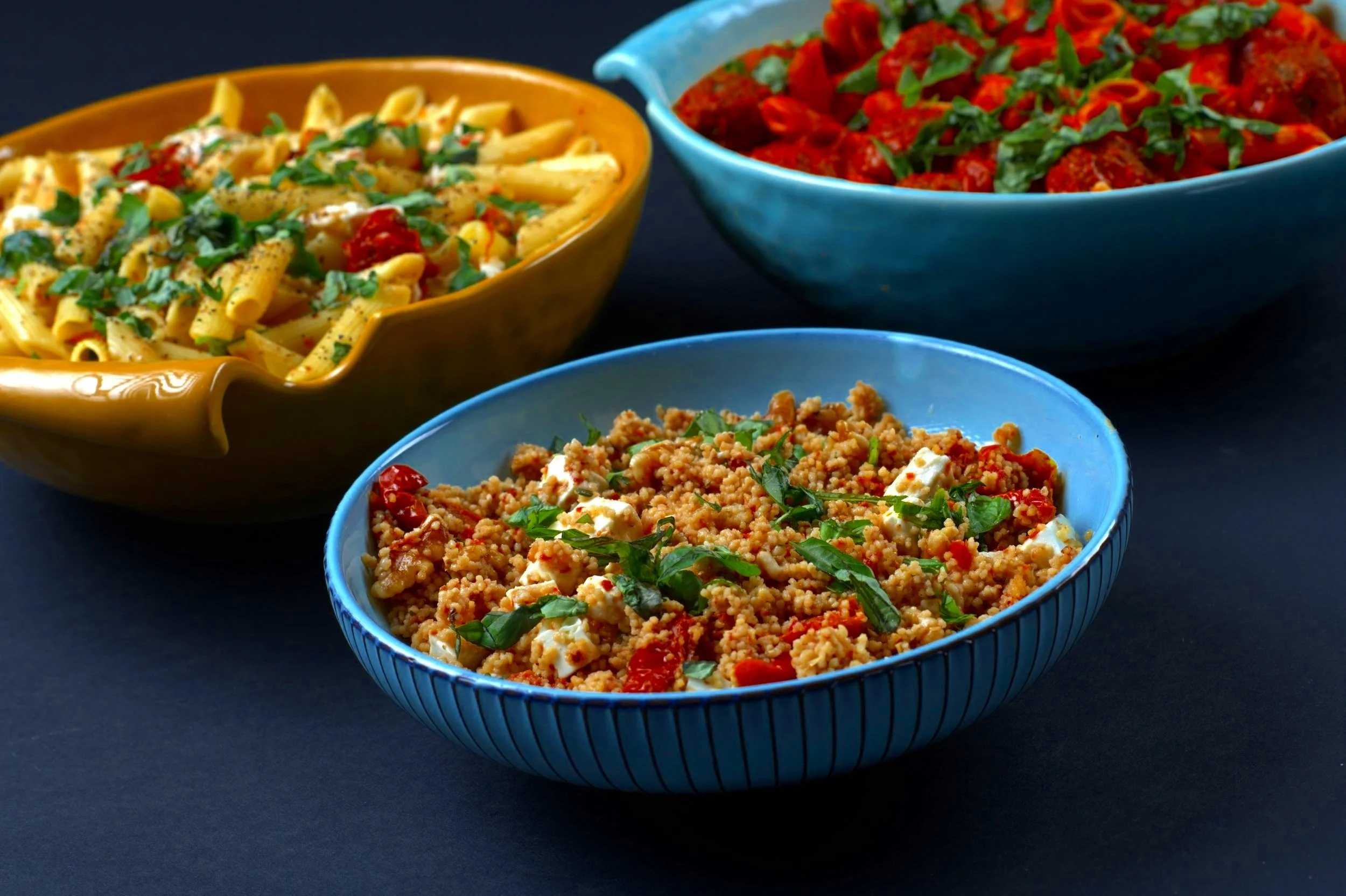 Three bowls with different dishes: pasta with tomatoes and herbs, couscous with feta and vegetables, and tortellini with tomato sauce.