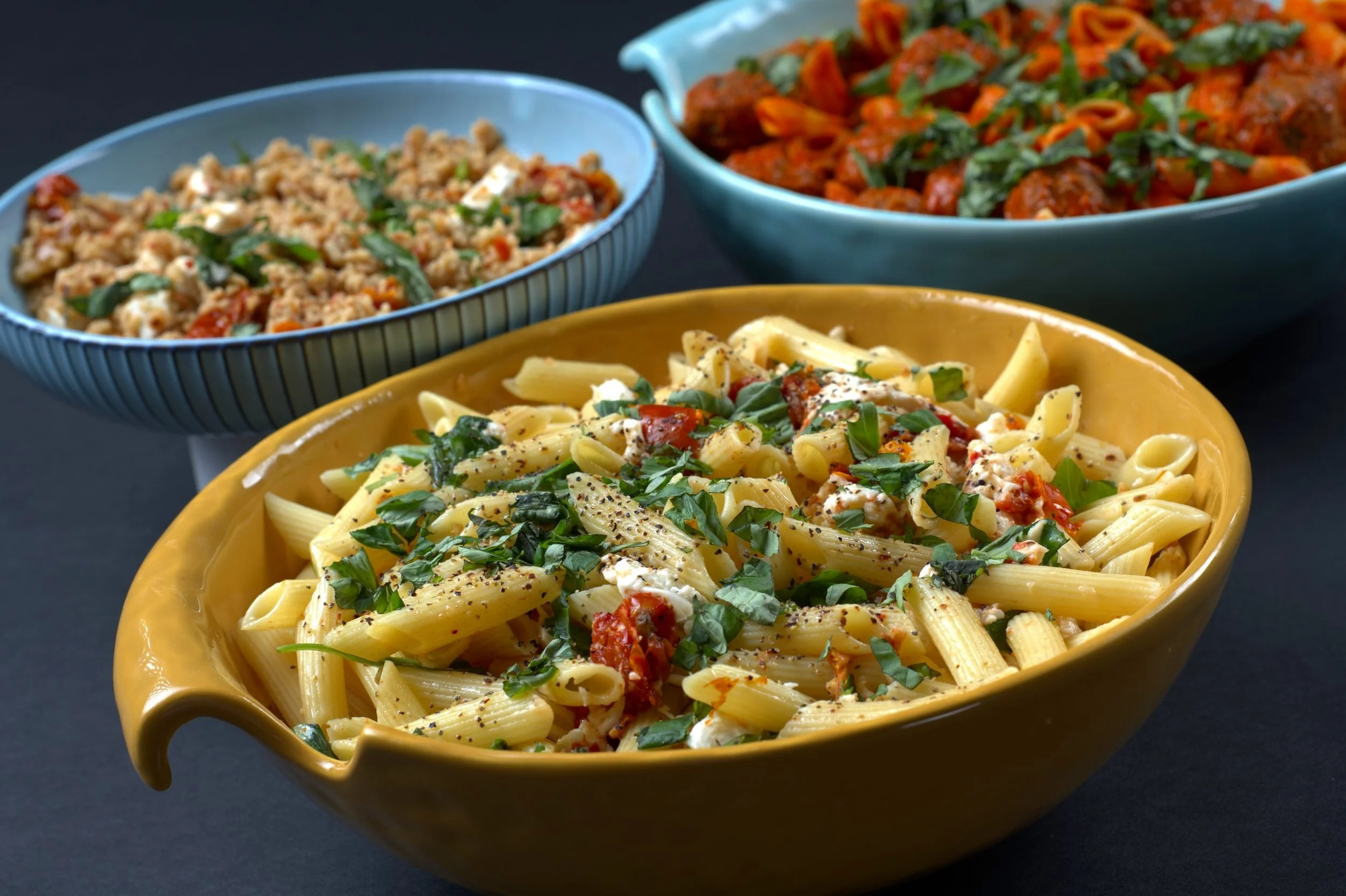 Three bowls of pasta dishes, featuring penne with herbs, spaghetti with meatballs, and a grain-based salad.