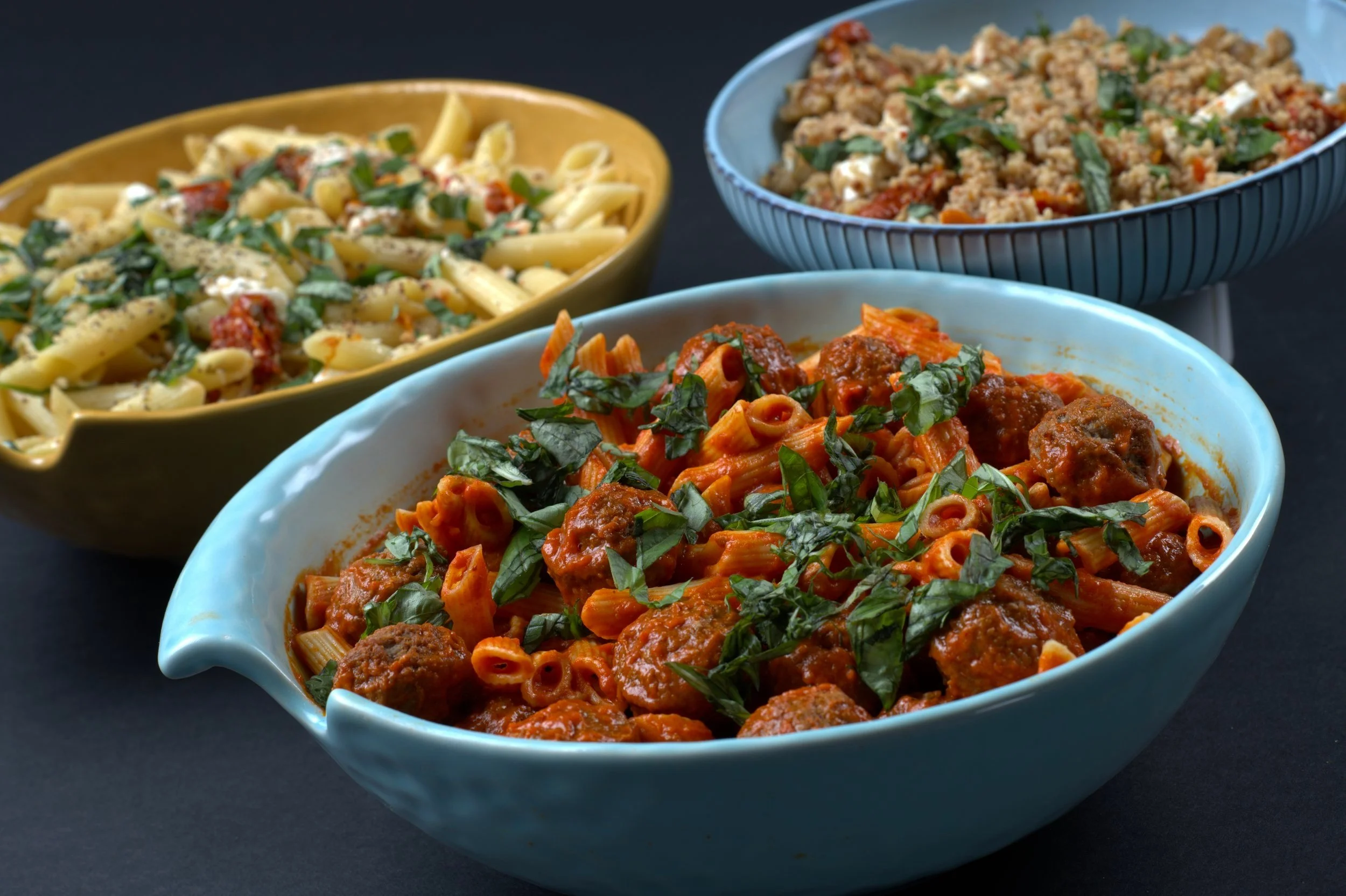 Assorted pasta dishes in serving bowls, including penne with meatballs and tomato sauce, pasta with cheese and herbs, and grain salad with vegetables.