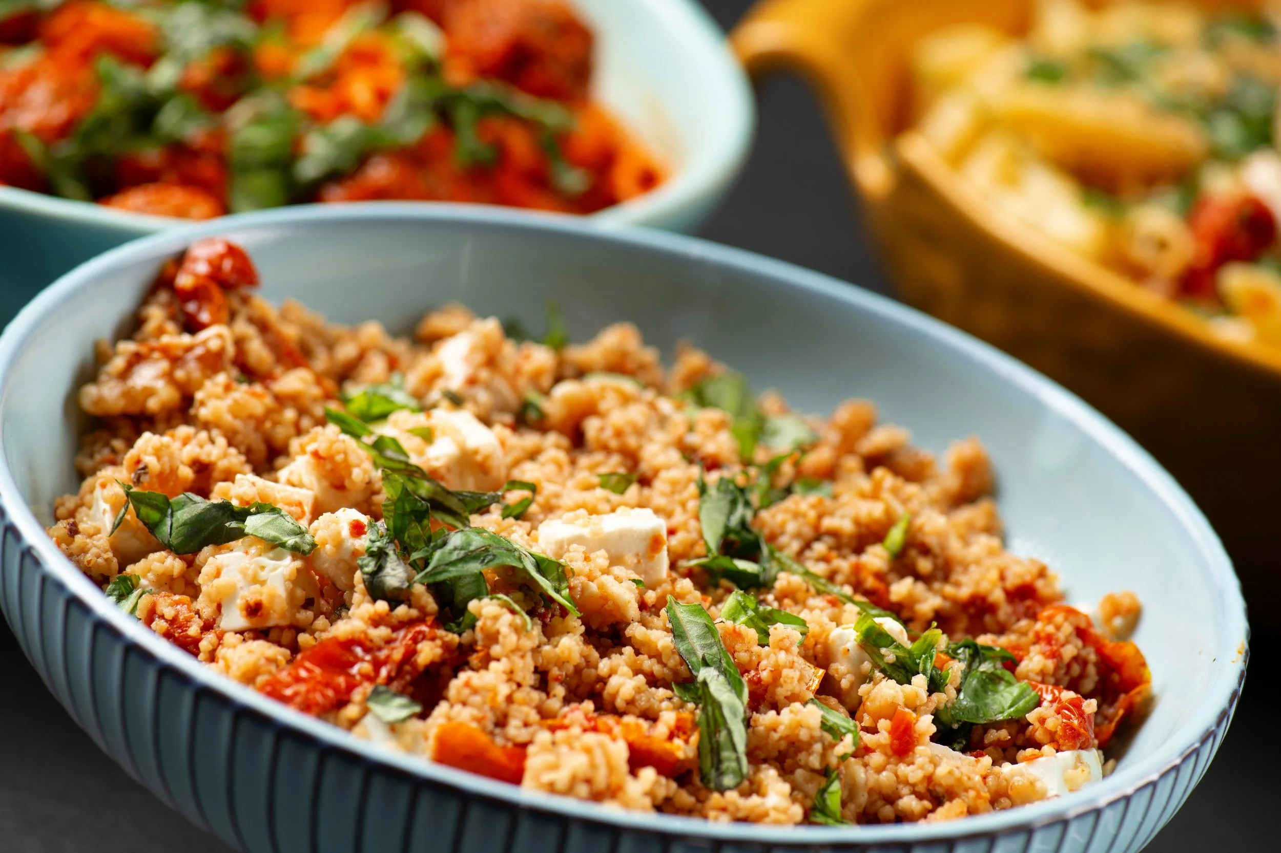 Close-up of a bowl of couscous salad with feta cheese, sun-dried tomatoes, and fresh herbs, with blurred dishes in the background.