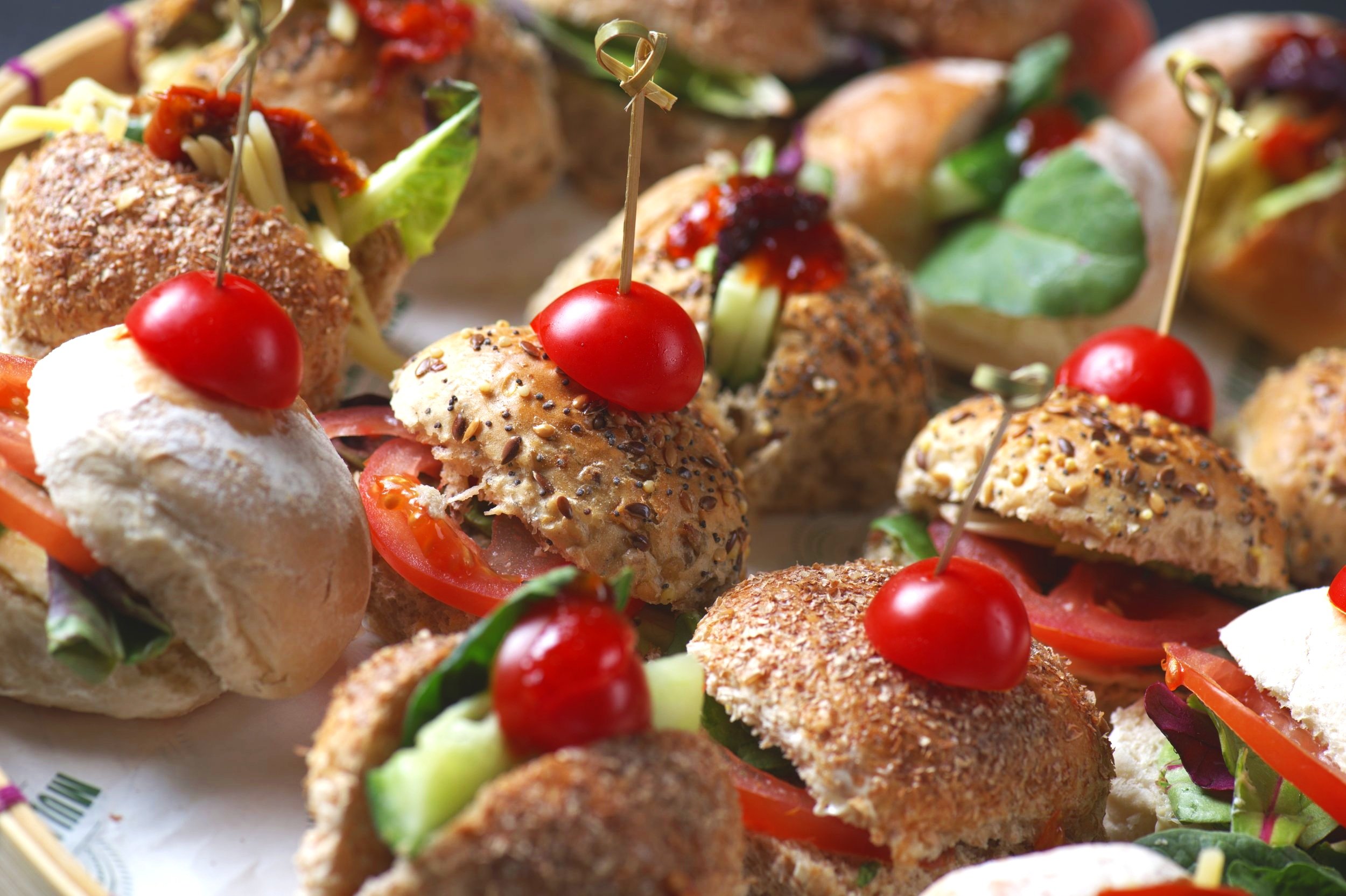 Assorted mini sandwiches with cherry tomatoes on top, featuring whole grain rolls, lettuce, cucumber, and tomato slices on a platter.