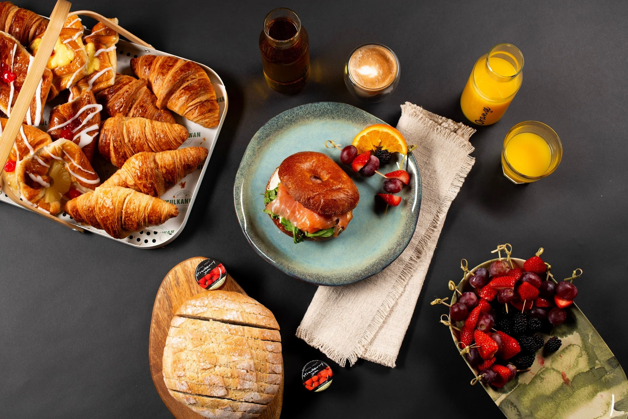 Assorted breakfast spread with croissants, bagel sandwich, fruits, bread, and drinks on a dark background.