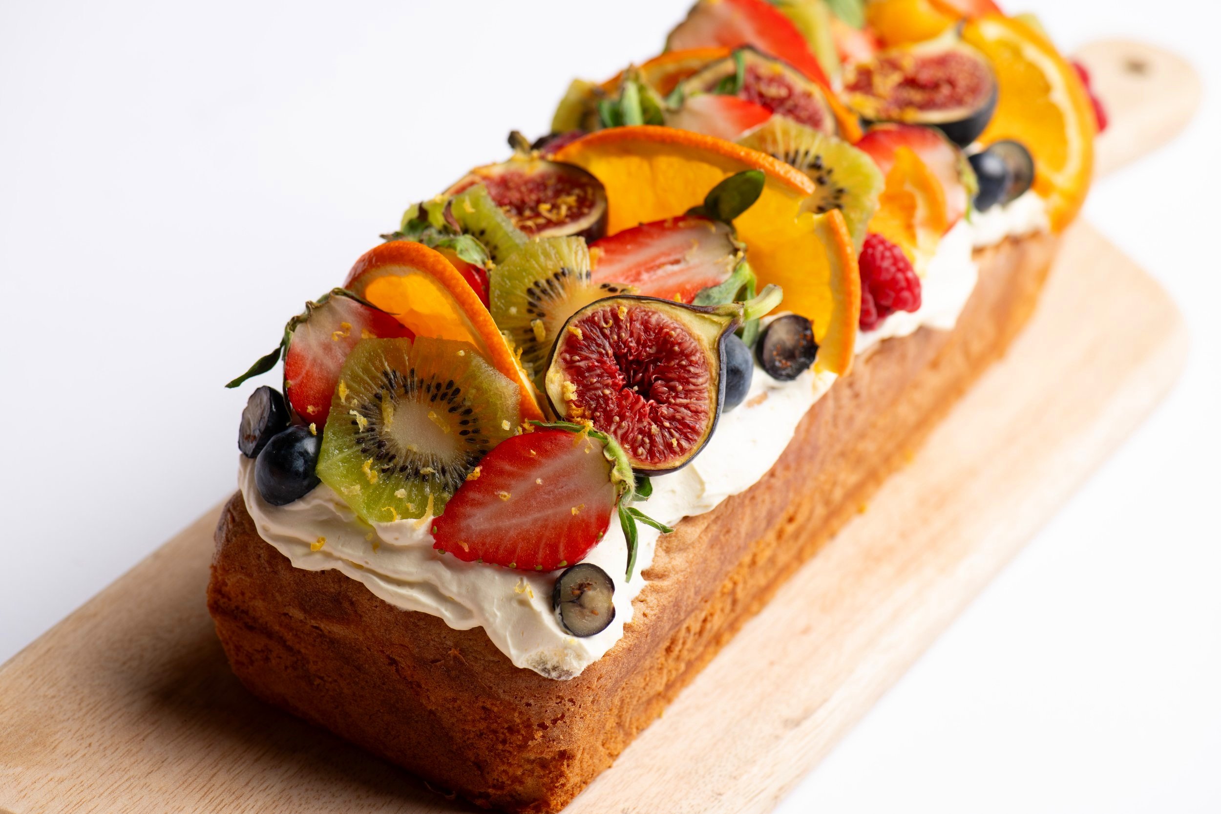 Fruit-topped cake with cream frosting featuring slices of strawberries, kiwi, oranges, figs, and berries on a wooden board.