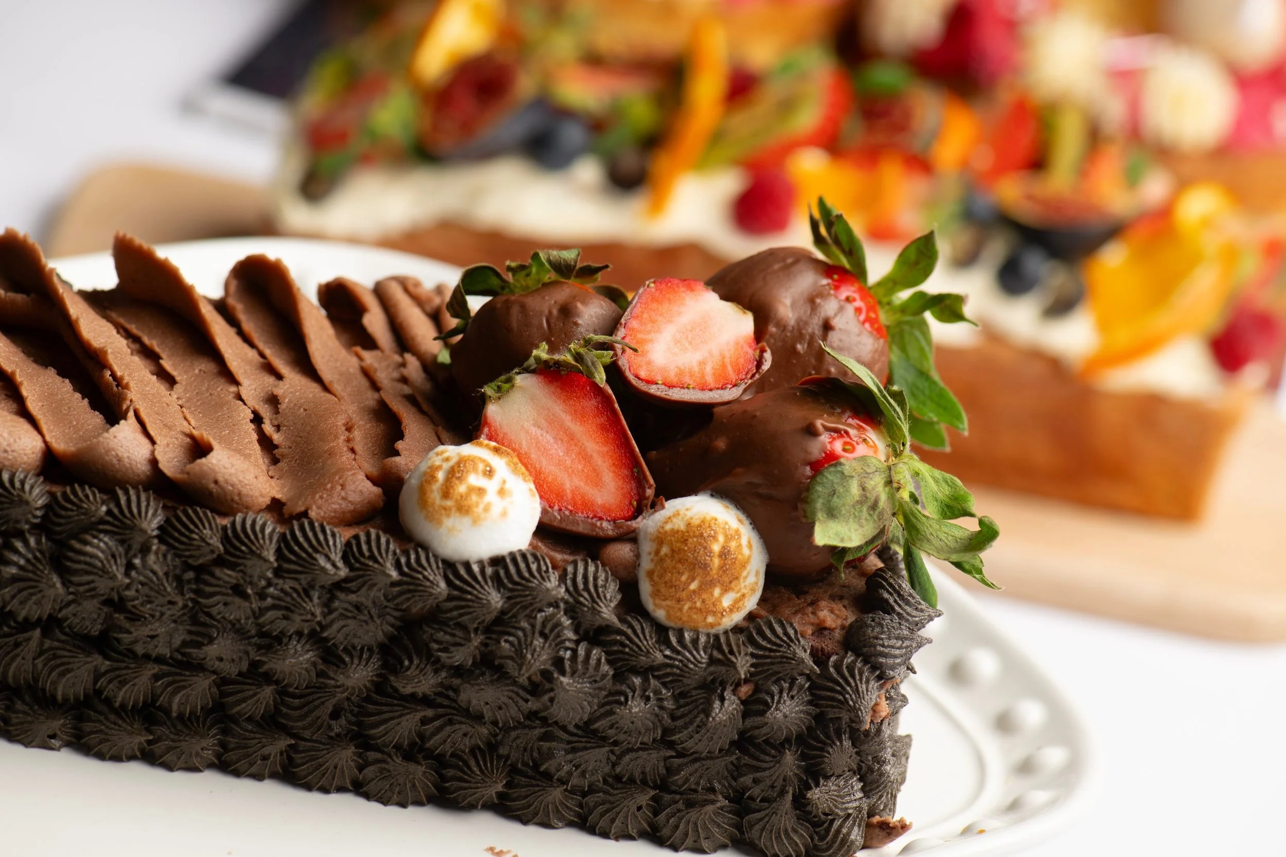 Close-up of a chocolate cake slice with chocolate frosting, topped with chocolate-covered strawberries and toasted marshmallows.