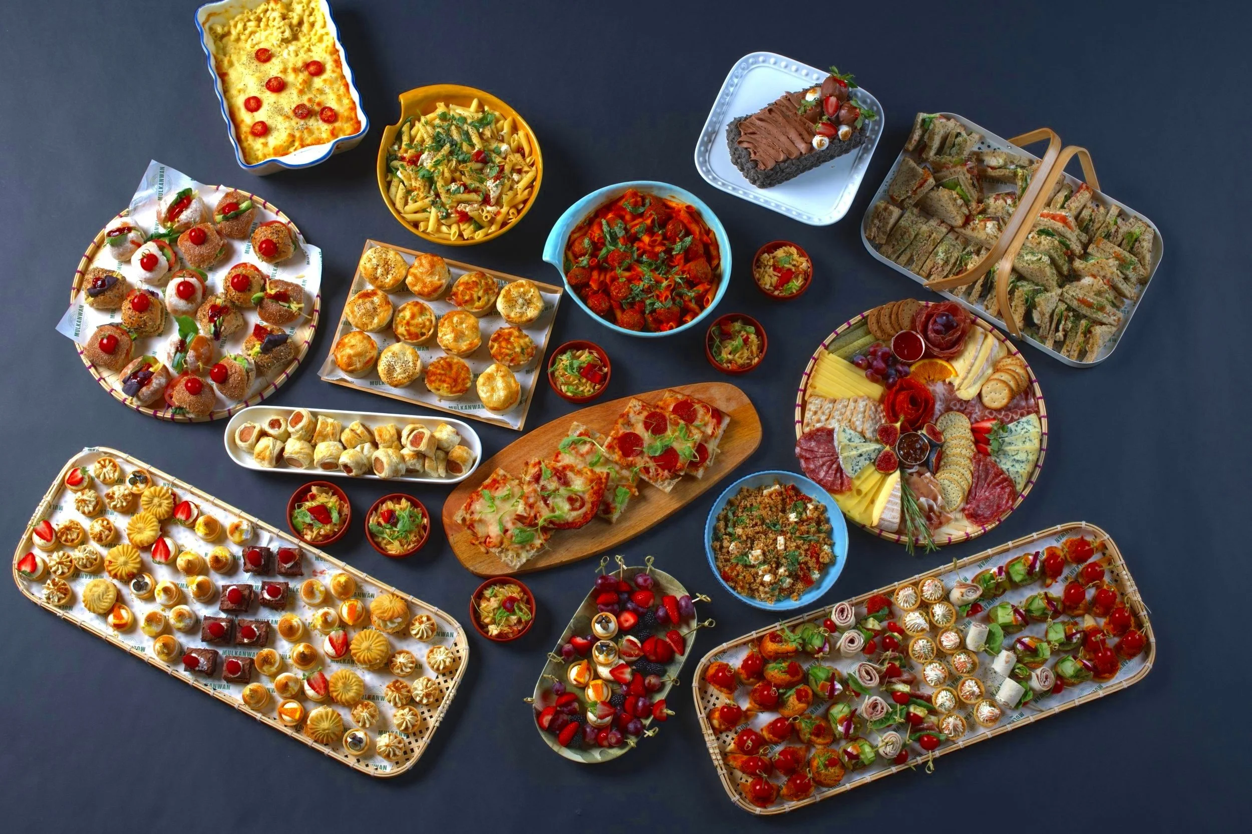 Assorted appetizers and pasta dishes including pizza, sandwiches, cheese platter, pasta salad, mini sausages, desserts, and salads on a dark background.