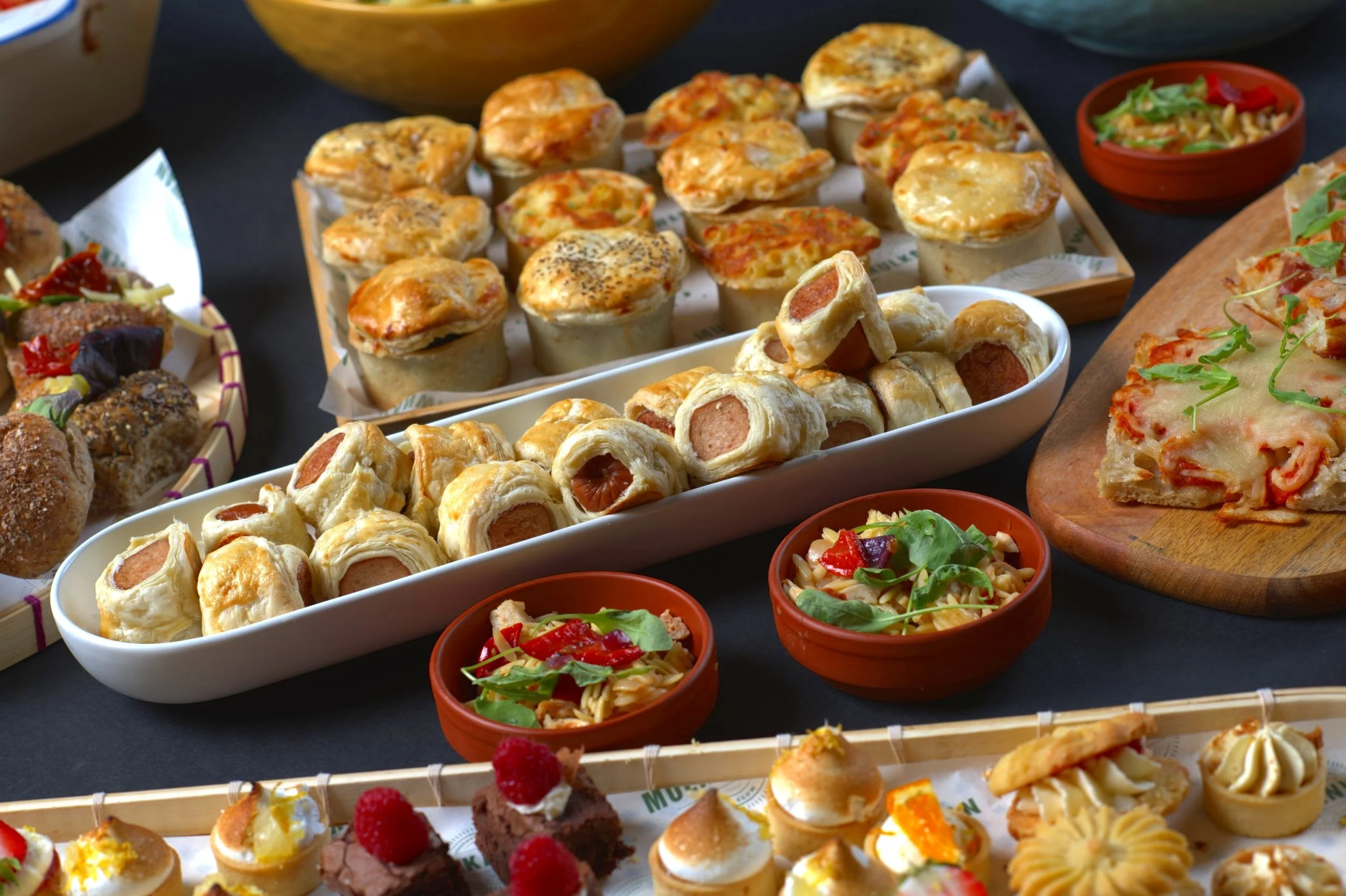 Assorted savory and sweet appetizers, including sausage rolls, mini pies, pizza slices, salads, and desserts on a table.