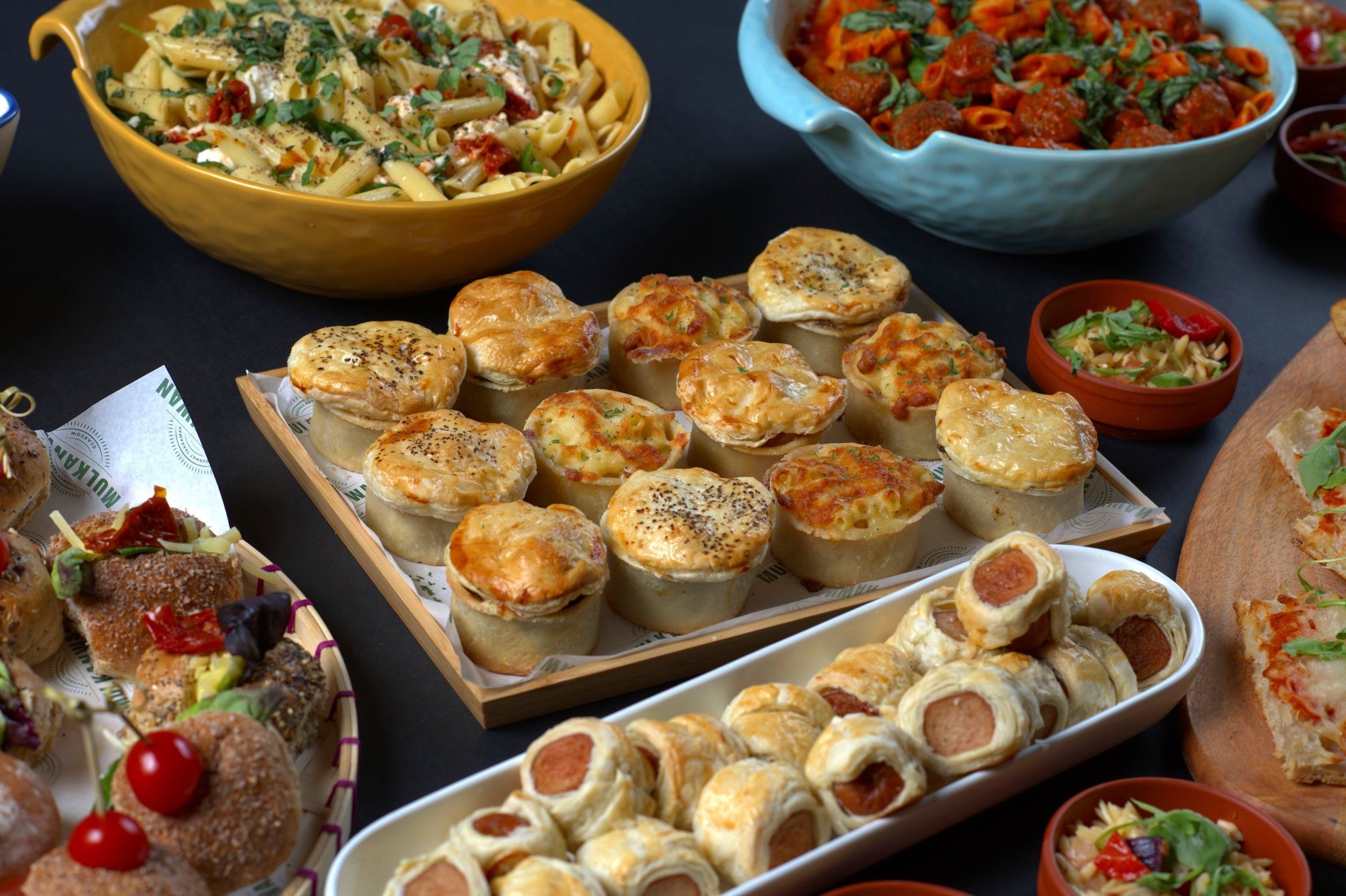 Assorted dishes including pasta, mini pies, sausage rolls, skewers, and salads on a table.