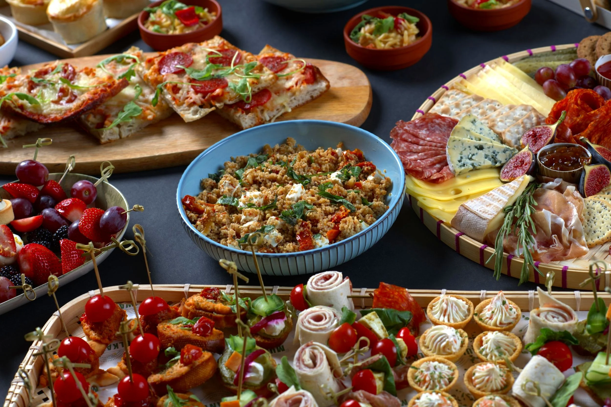 A table spread with assorted appetizers and snacks, including pizza slices on a wooden board, a bowl of couscous salad, a charcuterie board with cheeses, meats, figs, and crackers, a bowl of mixed fruit skewers, and various small appetizers like roll