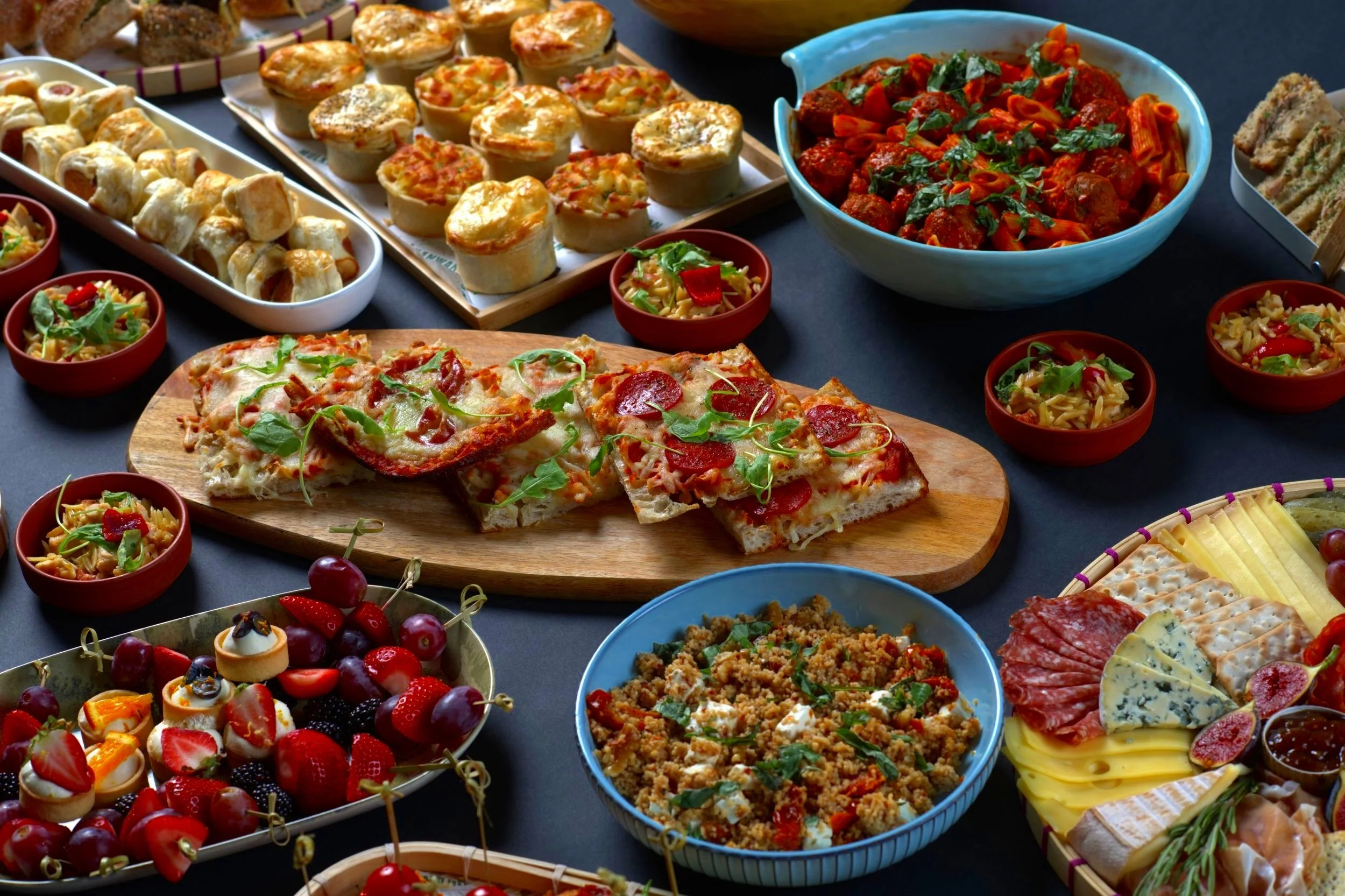 A variety of foods including pizza, mini pies, pasta with meatballs, assorted desserts, antipasto platter with cured meats, cheeses, crackers, grapes, and figs.