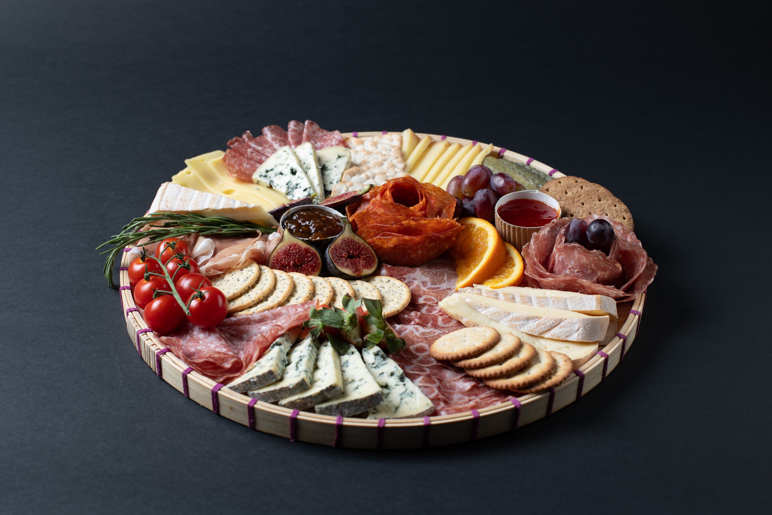 A charcuterie board featuring assorted cheeses, meats, crackers, figs, cherry tomatoes, orange slices, grapes, and a small cup of red sauce on a round wooden platter.