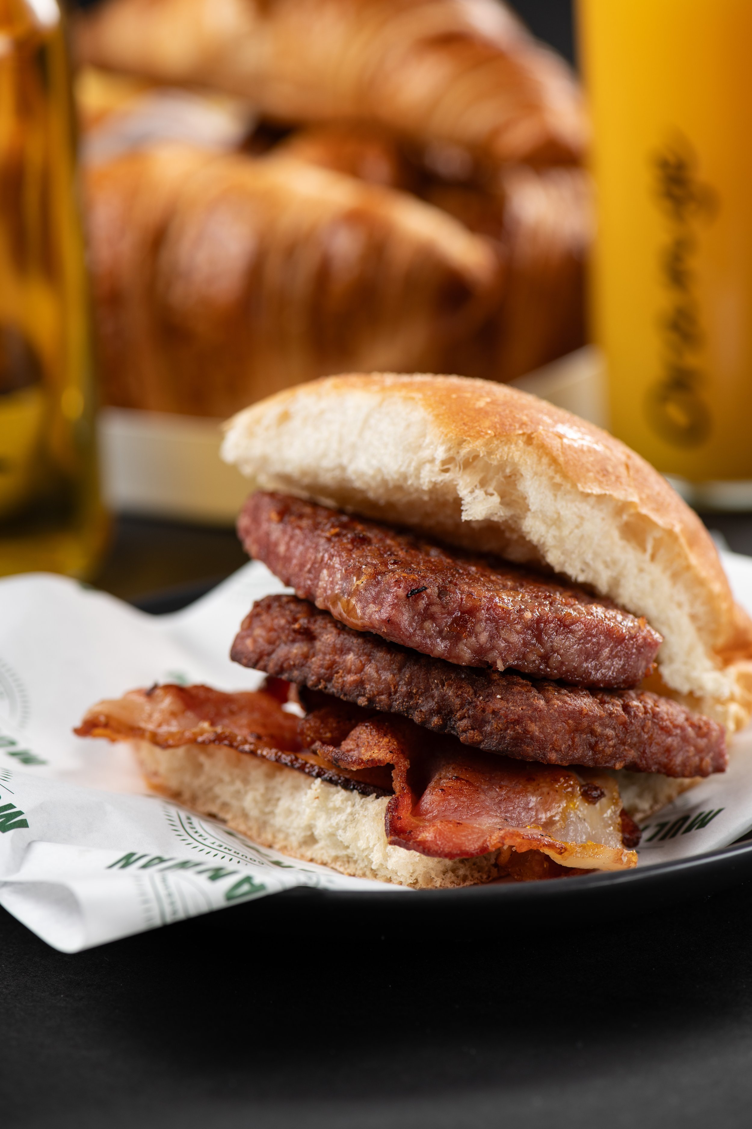 Burger with bacon on a plate, croissants and orange juice in the background.