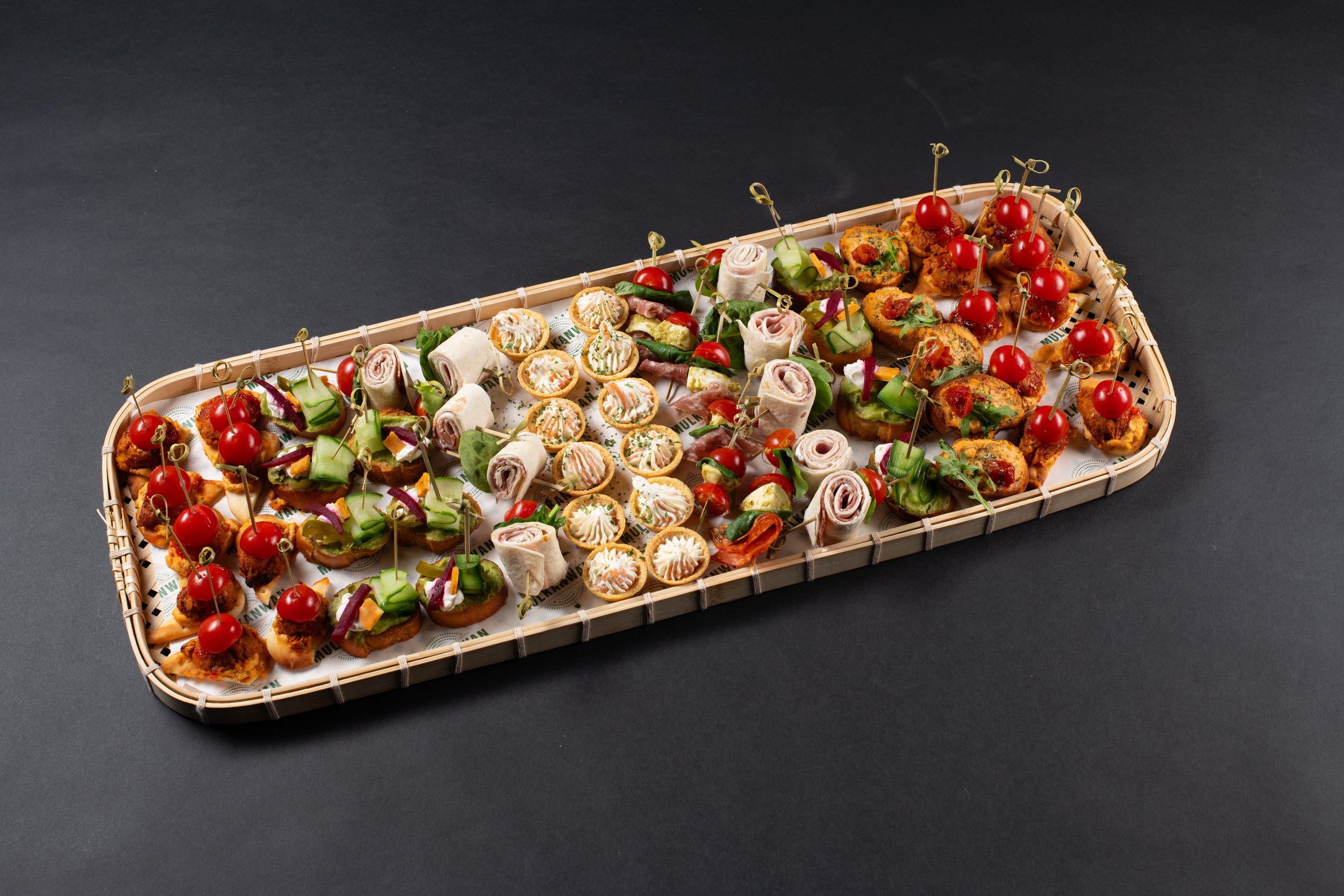 A platter of assorted appetizers on a dark background, featuring wraps, tartlets, skewered cherry tomatoes, and assorted finger foods arranged in rows.