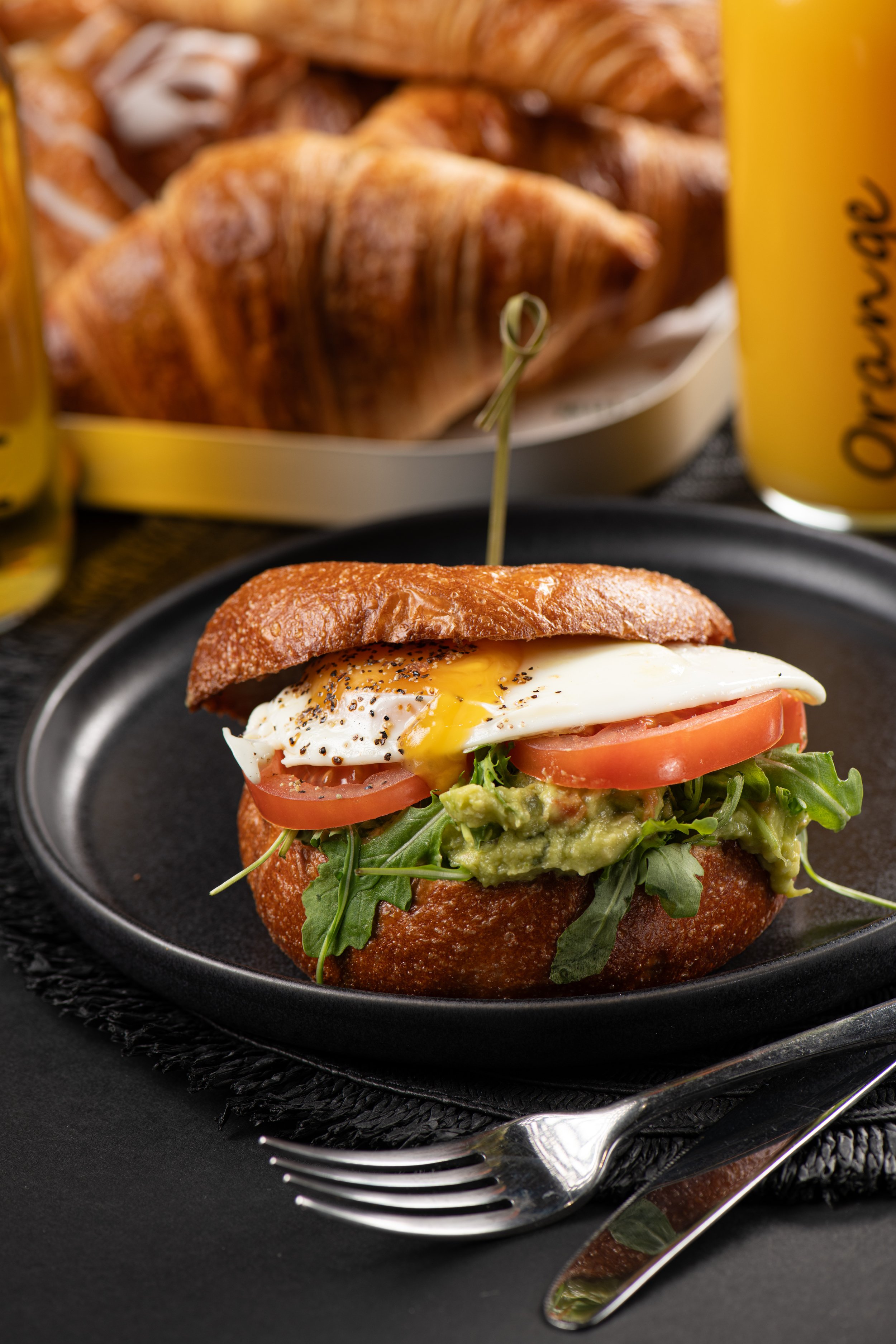 Breakfast sandwich with fried egg, tomato, avocado, and greens on a bun, served on a black plate with croissants and orange juice in the background.