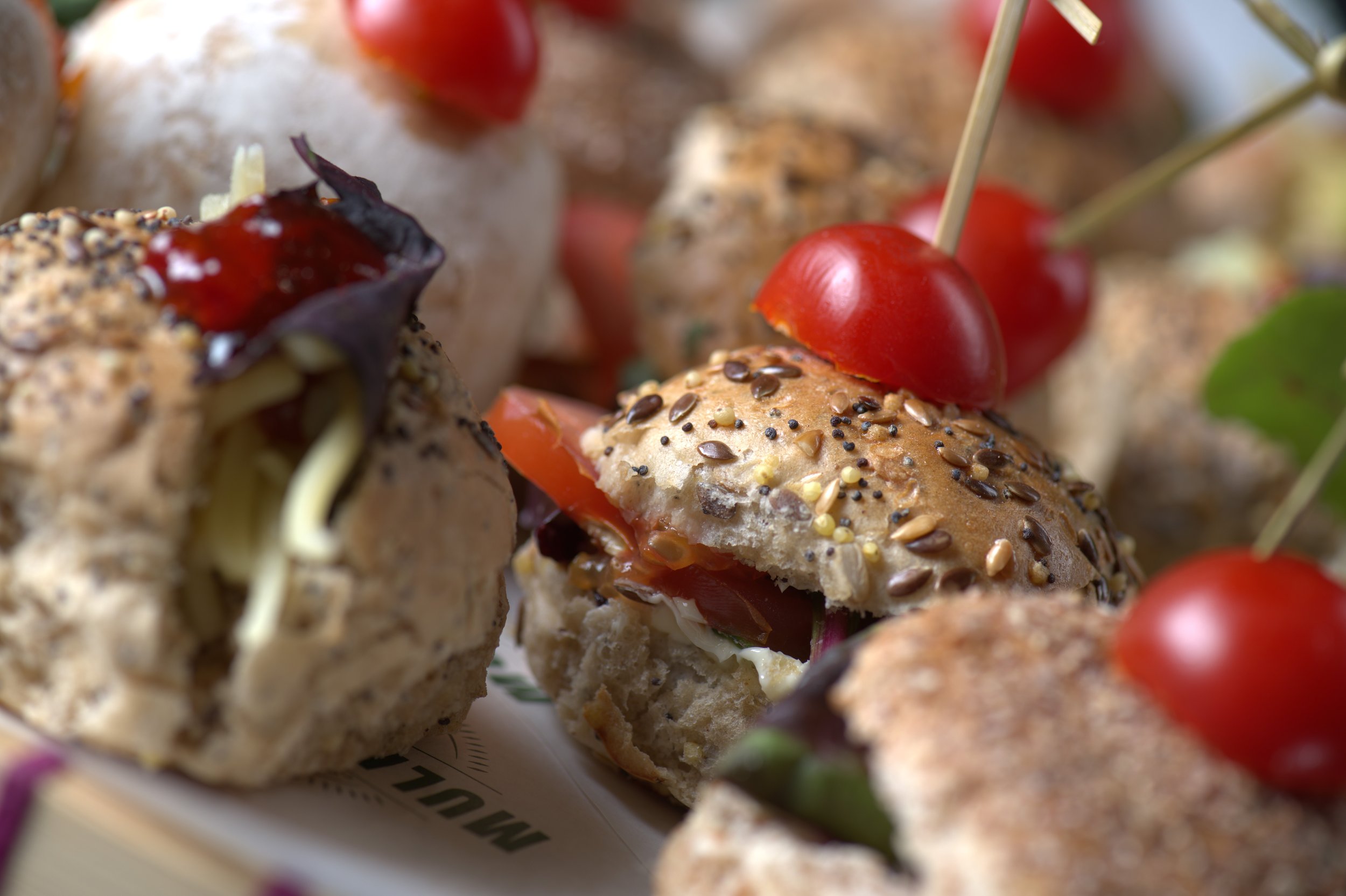 Close-up of assorted mini sandwiches with sesame and seed buns, garnished with cherry tomatoes and filled with vegetables and cheese.